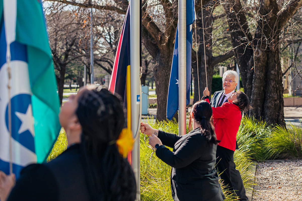 Professor Glyn Davis AC with 3 PM&C staff at flagpoles raising the Australian Aboriginal Flag, Torres Strait Islander Flag and the Australian National Flag. Professor Glyn Davis AC with 3 PM&C staff at flagpoles raising the Australian Aboriginal Flag, Torres Strait Islander Flag and the Australian National Flag.