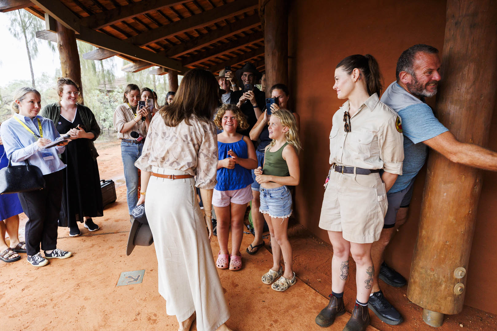 A group of people stand outdoors under a covered area, some taking photos of Her Majesty The Queen of Denmark speaking with two young girls stand smiling in the center. A woman in a khaki uniform looks on. A man leans against a wooden pillar.