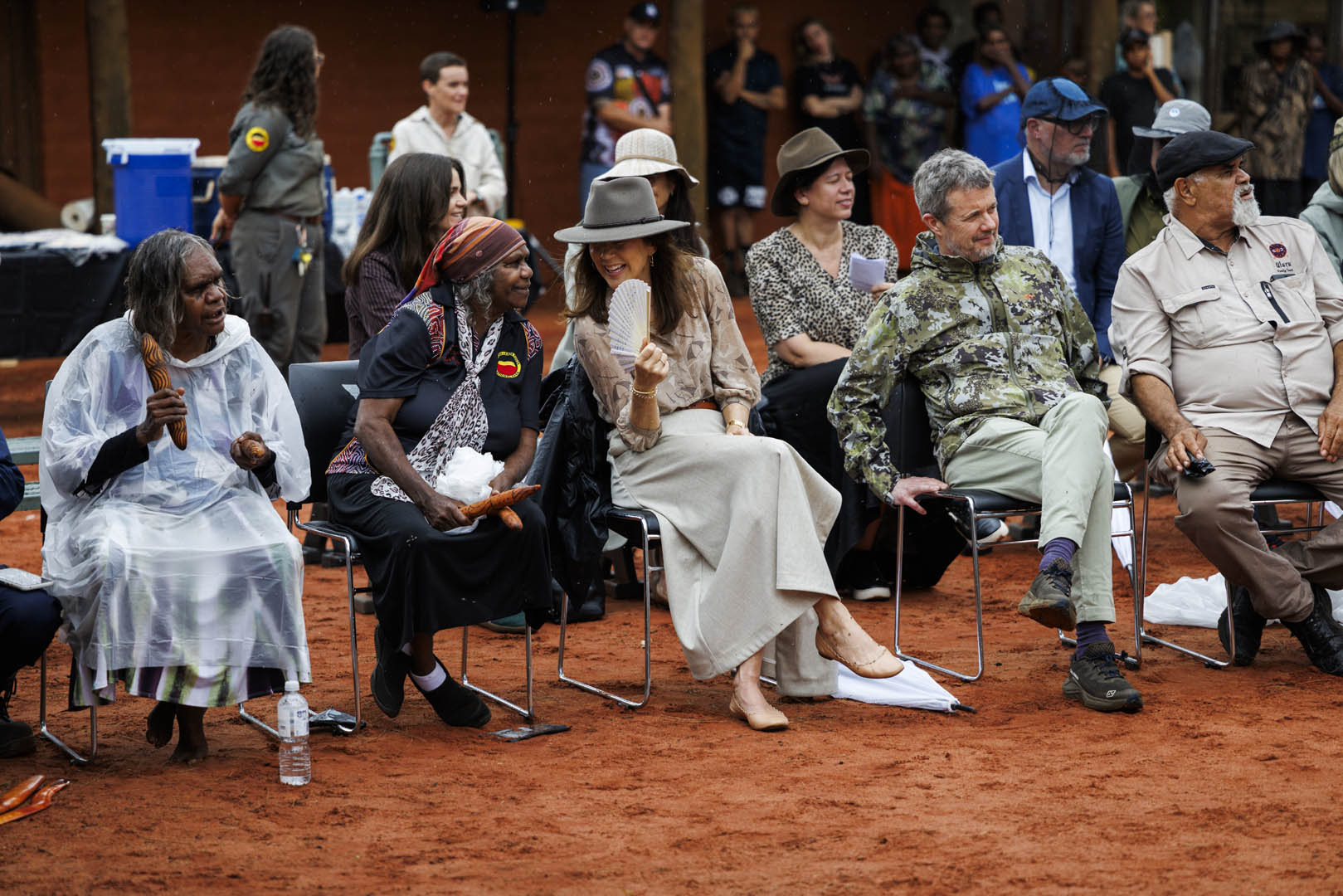 Outdoor shot of a diverse group seated on chairs, including Indigenous elders and Their Majesties The King and Queen of Denmark, during an event on a red-dirt ground with a crowd in the background.