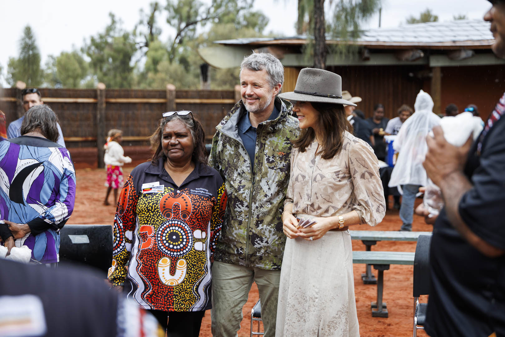 Their Majesties The King and Queen of Denmark meet with an indigenous woman. The woman is wearing a black shirt with a colourful design. His Majesty The King of Denmark is wearing a camo jacket and khaki pants, and Her Majesty The Queen of Denmark is wearing a hat and neutral coloured skirt and blouse.