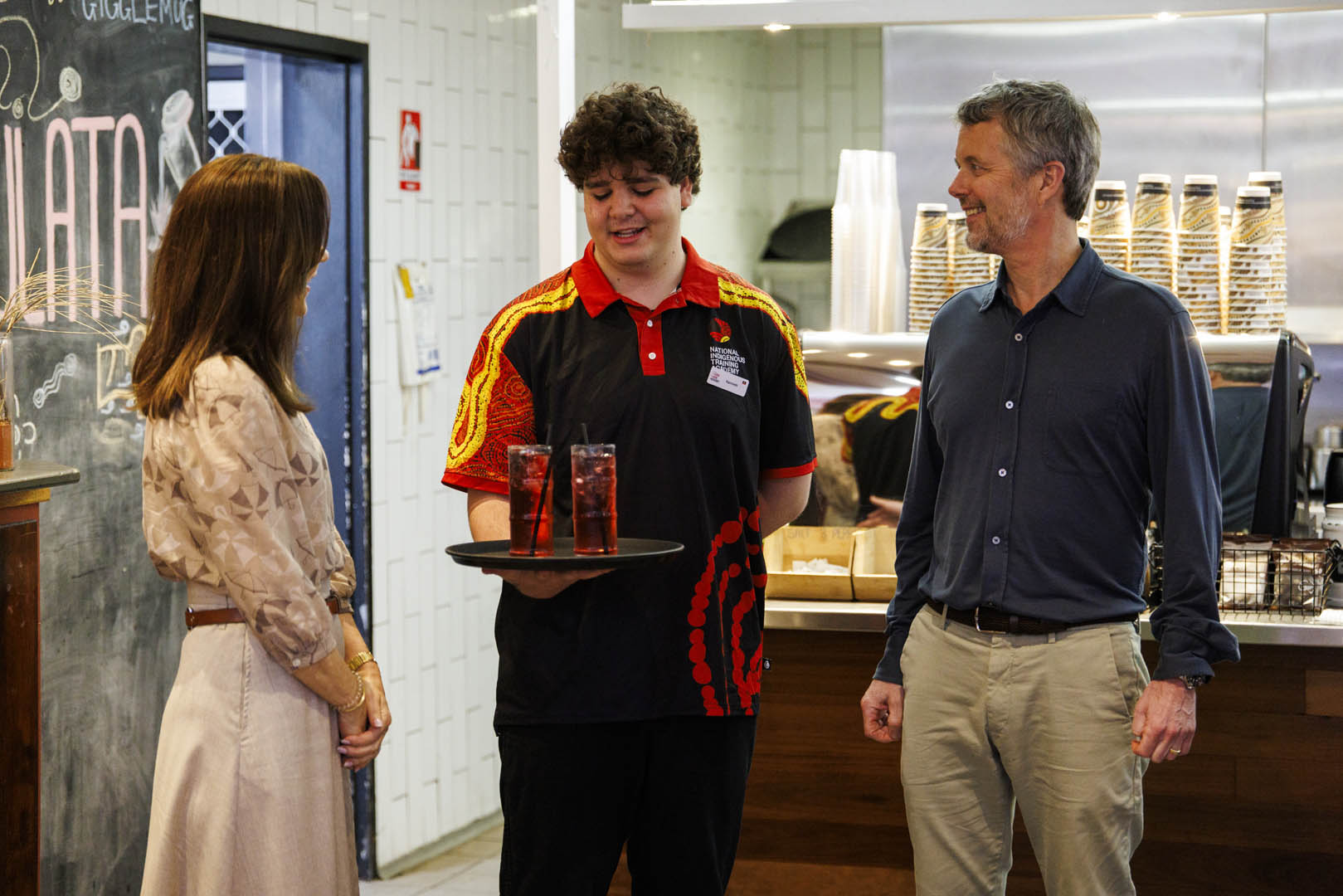 In a cafe, a student holding a tray with drinks speaks to Her Majesty The Queen of Denmark as His Majesty looks on. The server wears a black shirt with red and yellow patterns. "NATIONAL INDIGENOUS TRAINING ACADEMY" is visible on the shirt. 
