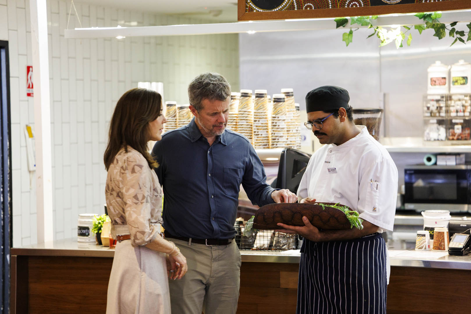 Their Majesties The King and Queen of Denmark admire a dish of green vegetables held by a chef in a kitchen. The chef wears a white uniform, apron, and hat. 