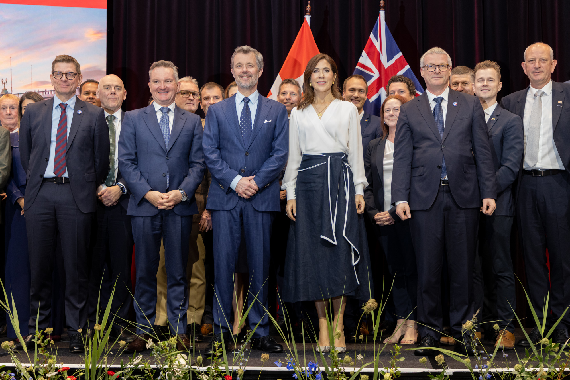 Group poses before flags with Their Majesties The King and Queen of Denmark. Both smile in front of a crowd. His Majesty wears a blue suit and tie, whileHer Majesty wears a white wrap top and navy skirt.