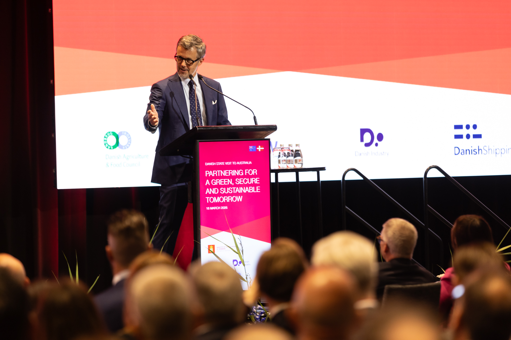 His Majesty The King of Denmark speaks at a podium labeled "DANISH STATE VISIT TO AUSTRALIA PARTNERING FOR A GREEN, SECURE AND SUSTAINABLE TOMORROW 18 MARCH 2026" before an audience. Logos for Danish Agriculture & Food Council, Danish Industry, and Danish Shipping are displayed.