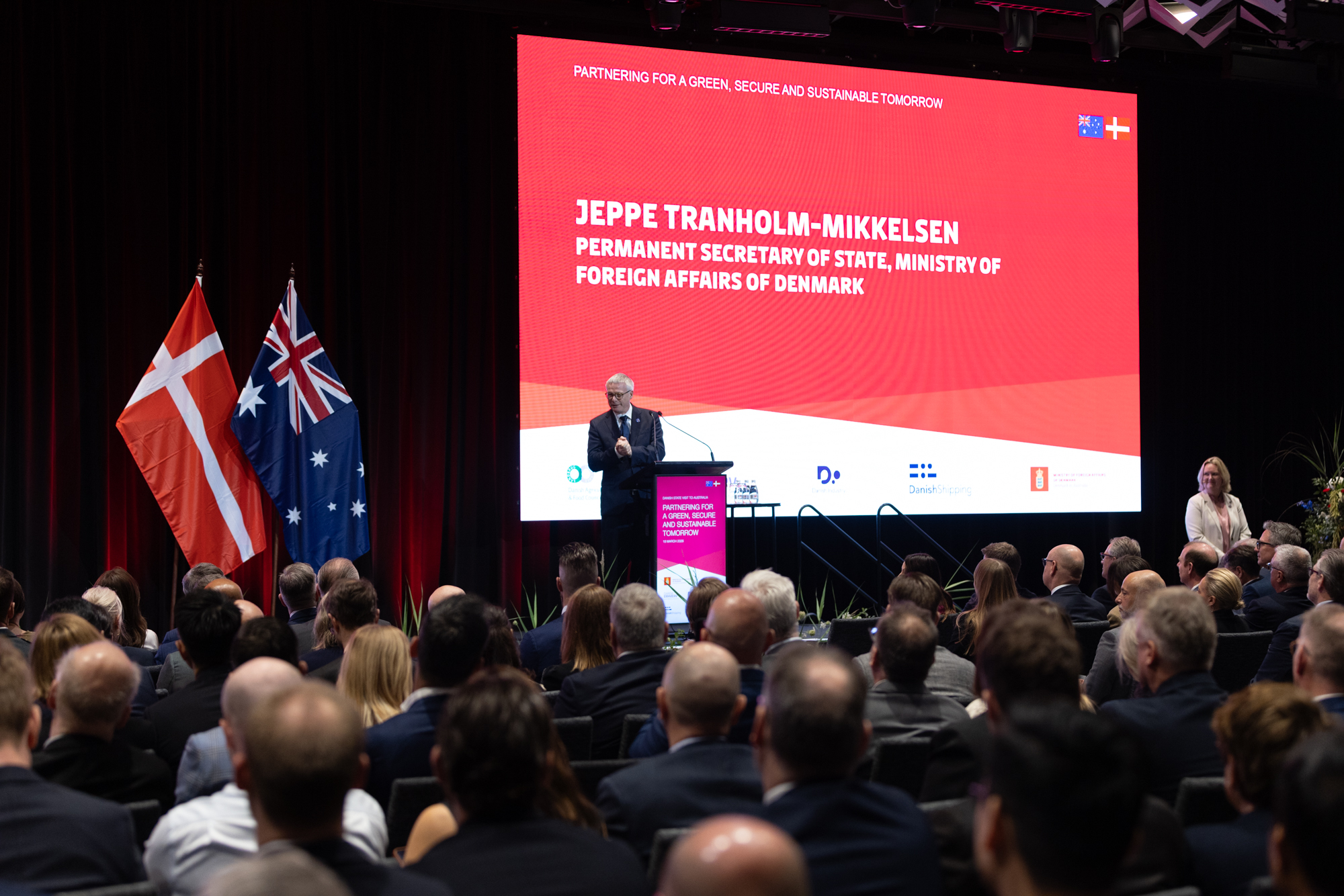 A speaker addresses an audience at a conference with Danish and Australian flags displayed. A screen reads: "PARTNERING FOR A GREEN, SECURE AND SUSTAINABLE TOMORROW JEPPE TRANHOLM-MIKKELSEN PERMANENT SECRETARY OF STATE..."