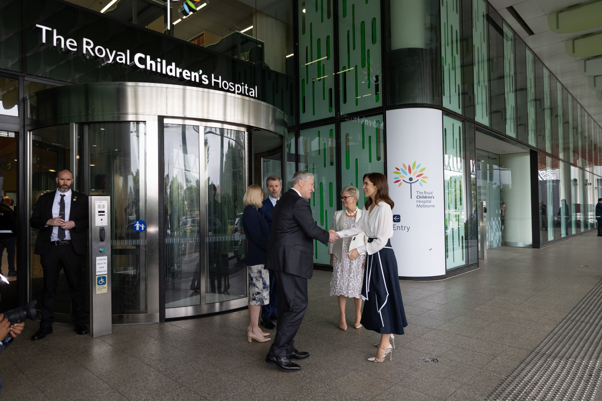 At 'The Royal Children's Hospital', Her Majesty The Queen of Denmark in a white top and navy skirt greets a man in a suit with a handshake. Other people stand nearby outside the 'Entry'. A sign reads: 'The Royal Children's Hospital Melbourne'.