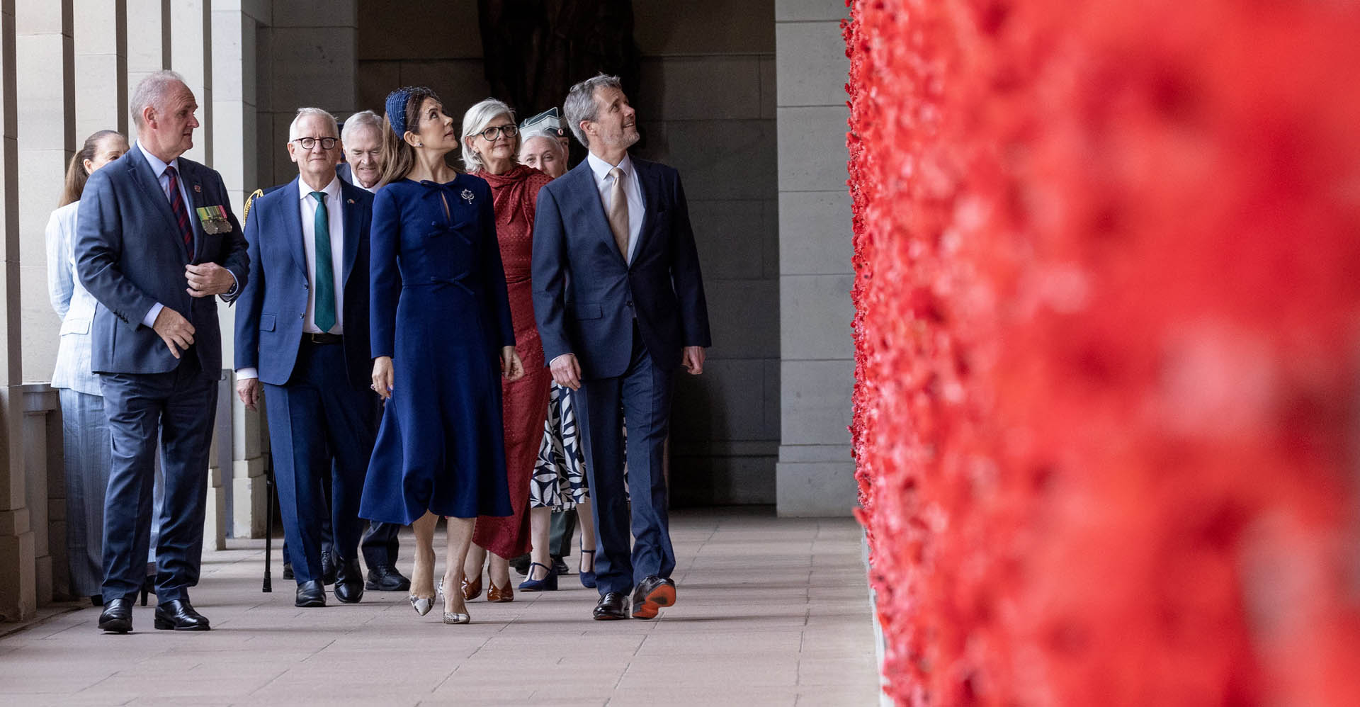 Group of people incuding Their Majesties The King and Queen of Denmark gaze upwards at the Roll of Honour at the Australian War Memorial. The group, consisting of men and women of varying ages, is dressed in suits and dresses. They stand on a tiled floor with columns in the background.
