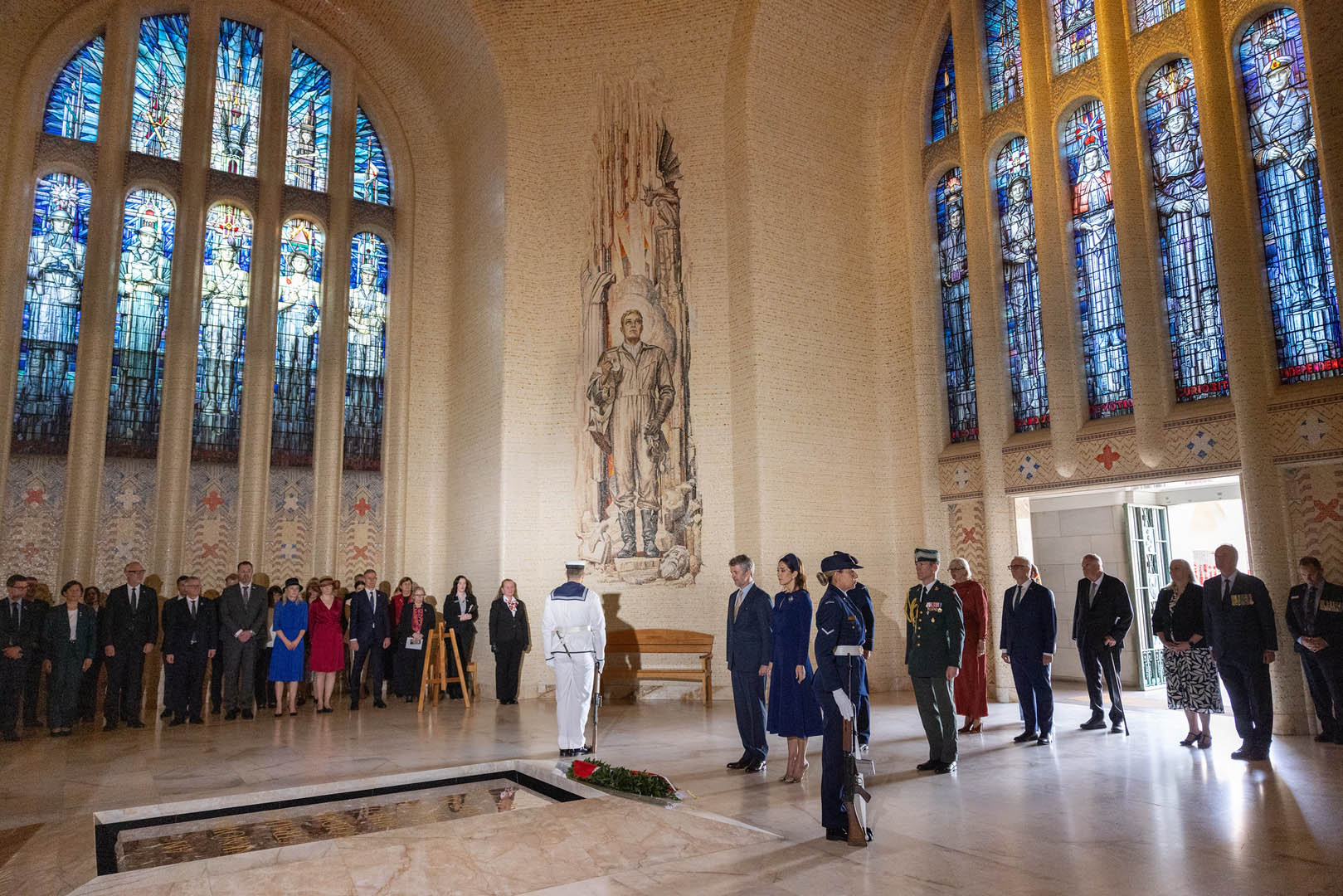 Interior view of the Hall of Memory at the Australian War Memorial in Canberra, people stand along the walls. The Hall features tall stained glass windows and a mosaic of an Australian serviceman. Their Majesties The King and Queen of Denmark stand in silence as they laid a wreath.