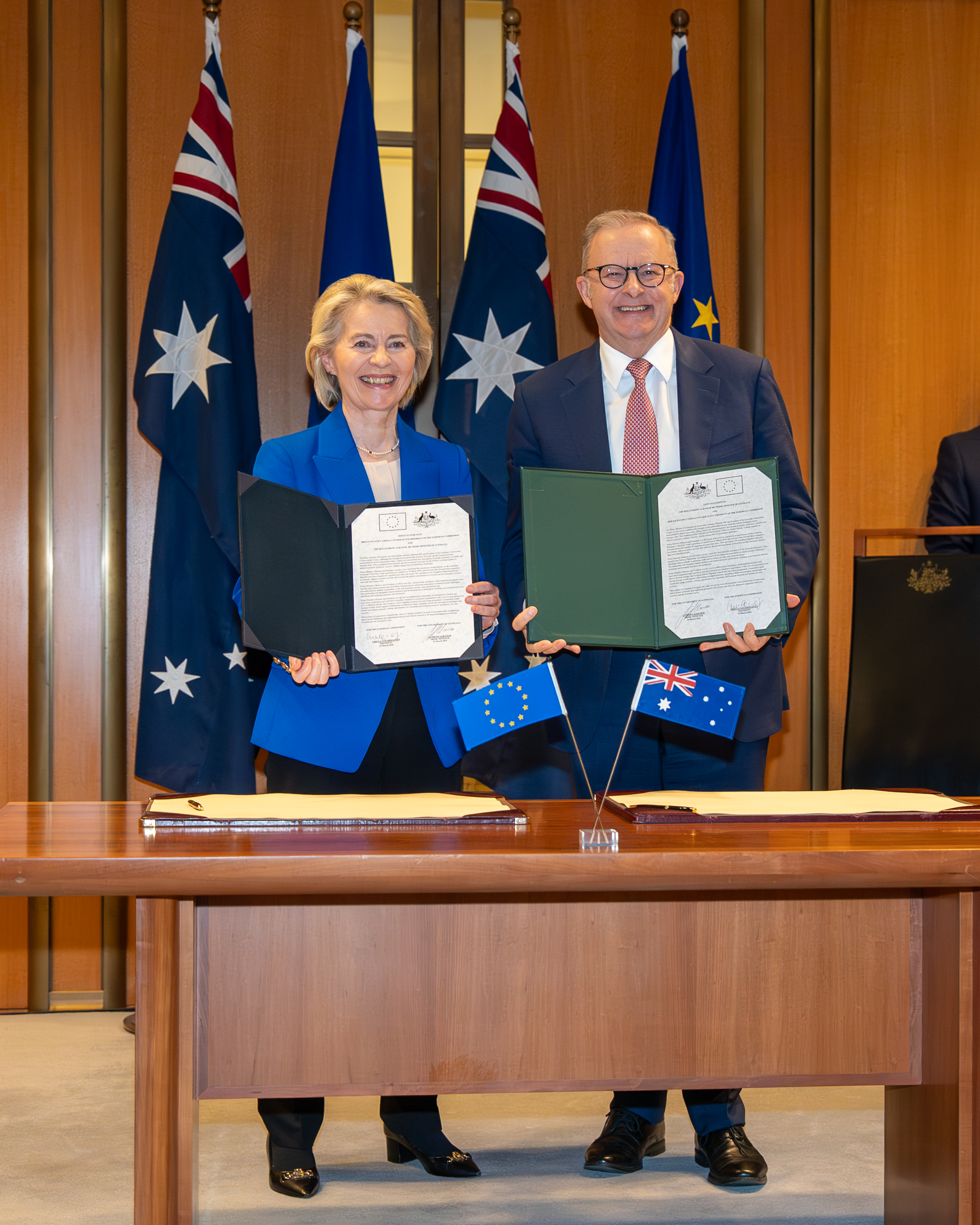 President von der Leyen in a blue blazer and Prime Minister Anthony Albanese in a suit stand behind a table holding signed documents, with Australian and European Union flags behind them.