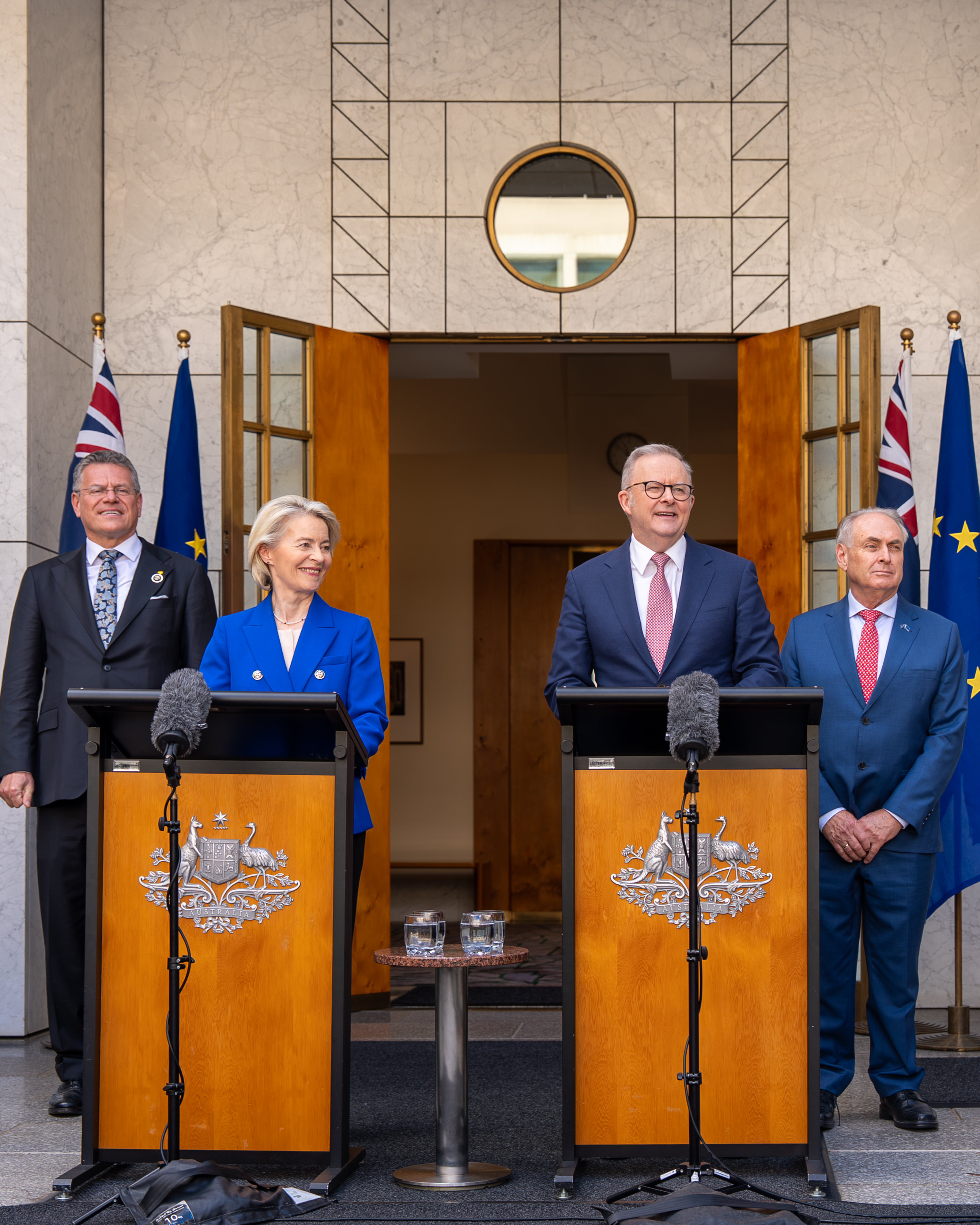 President von der Leyen and Prime Minister Albanese stand at podiums outdoors in front of a building entrance, with Australian and European Union flags behind them and officials standing nearby.