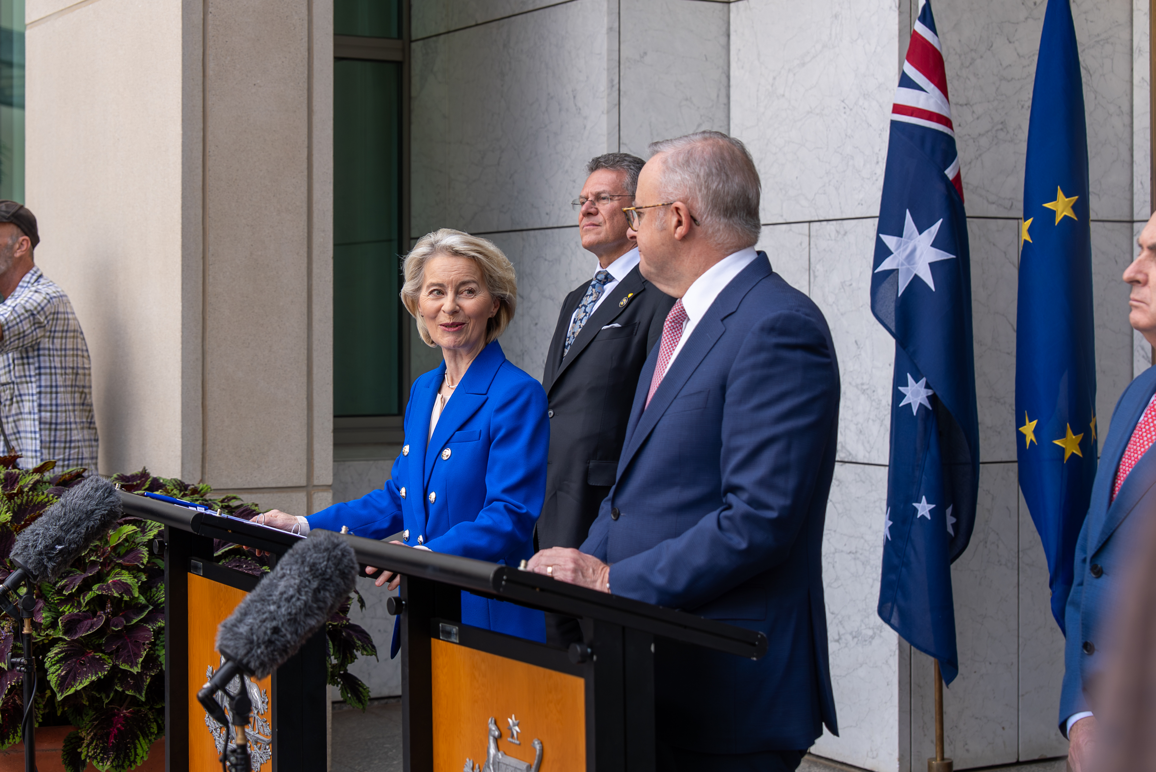 President von der Leyen in a blue blazer speaks at a podium outdoors while Prime Minister Albanese in a suit stands beside her, with microphones and flags visible.