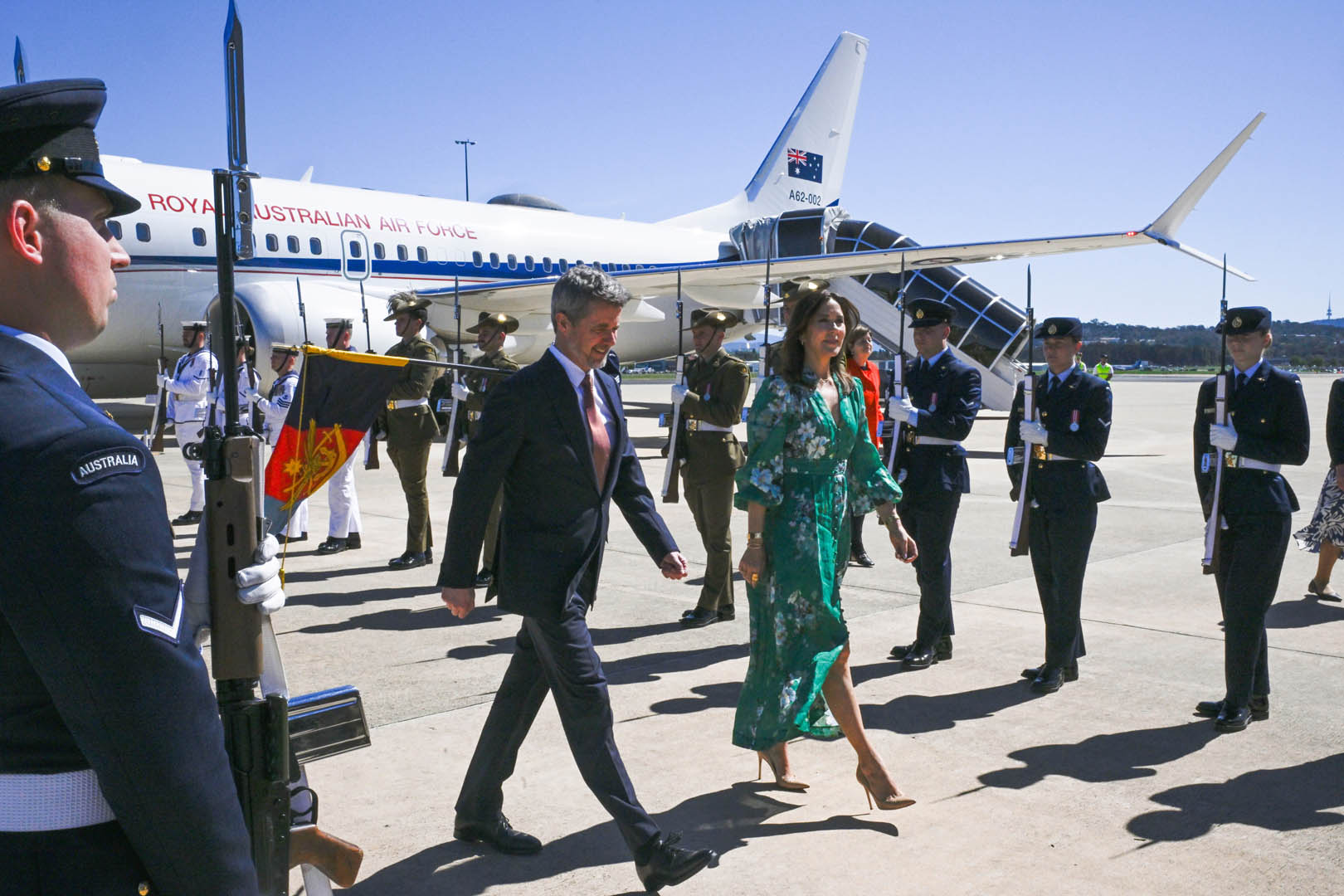 Their Majesties The King and Queen of Denmark are walking on the tarmac, with soldiers lining the path and a "ROYAL AUSTRALIAN AIR FORCE" plane in the background. The plane's tail has "A62-002" written on it. The soldiers are holding rifles with bayonets.