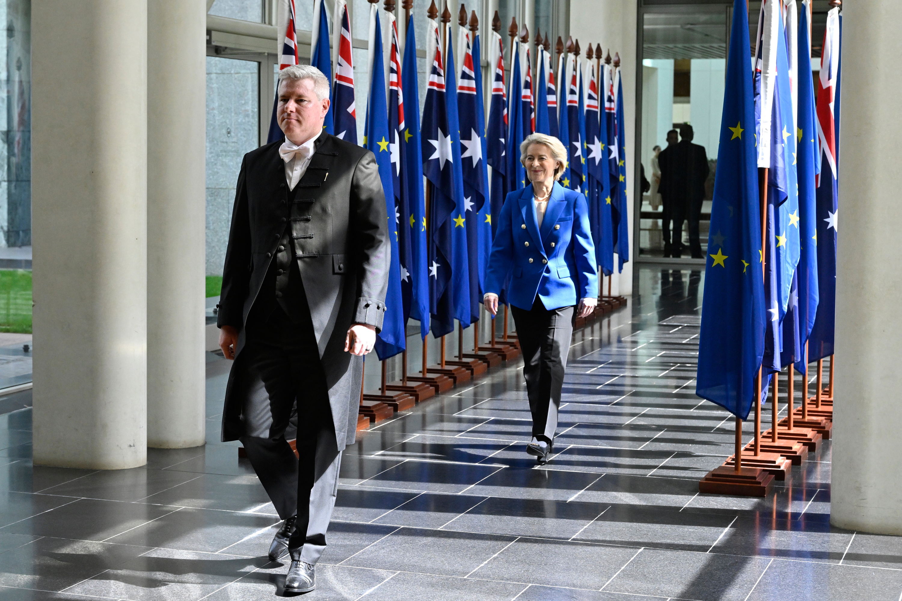 Ursula von der Leyen in a blue blazer walks through a corridor lined with Australian and European Union flags inside Parliament House, accompanied by an official.