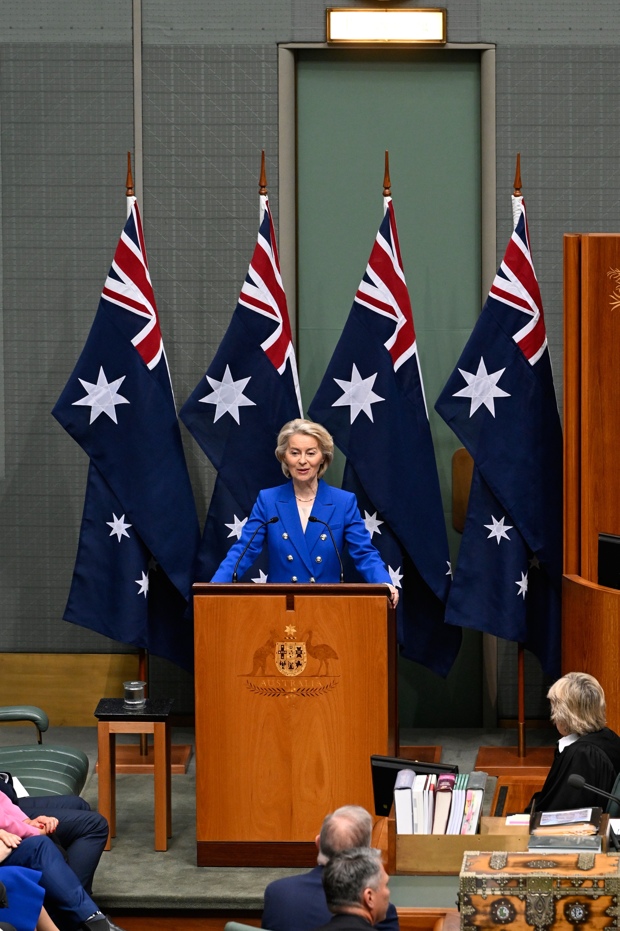 Ursula von der Leyen in a blue blazer stands at a wooden lectern in the House of Representatives, speaking in front of multiple Australian flags.