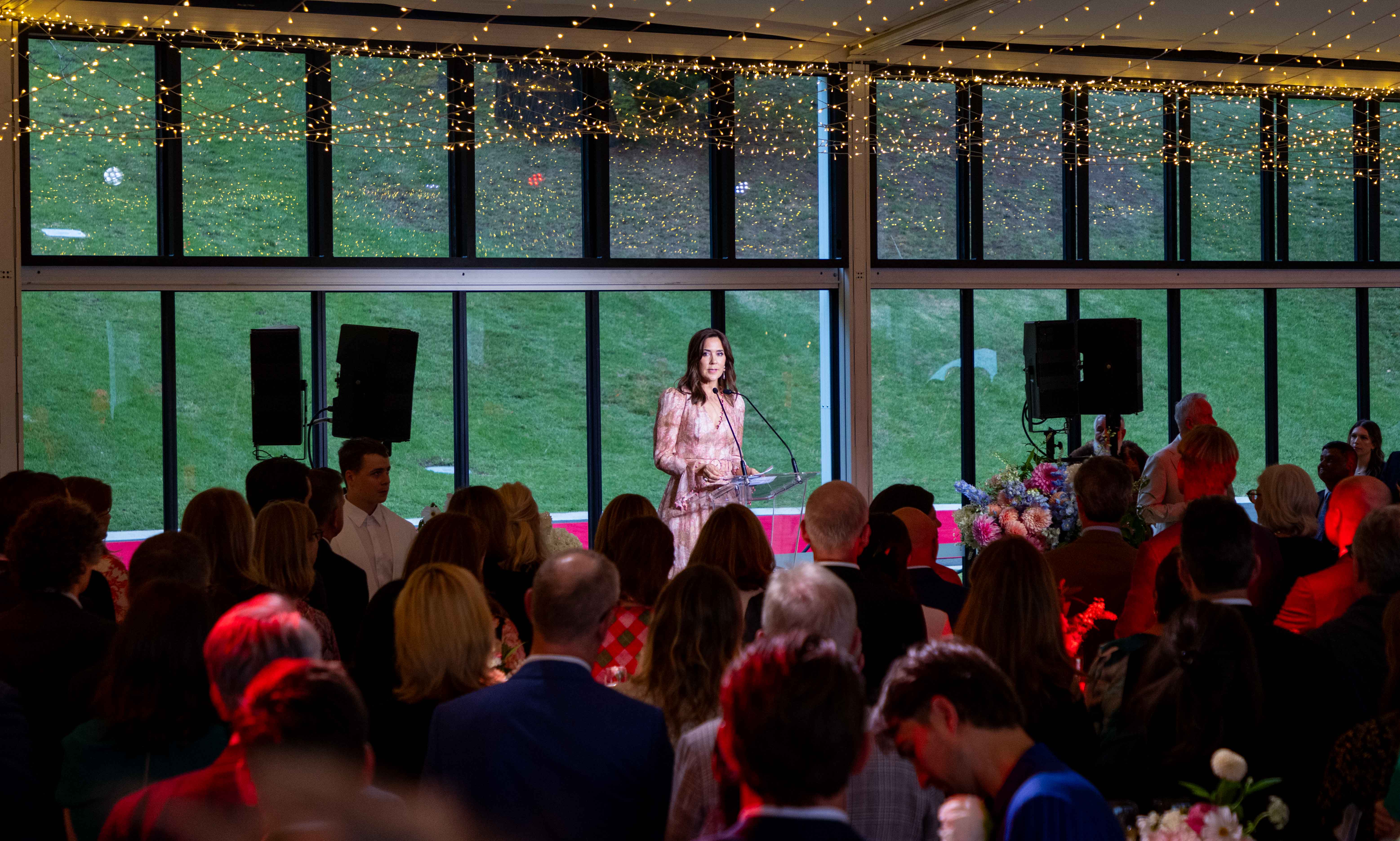 Her Majesty The Queen of Denmark speaks at a podium to a crowd at the Royal Botanic Gardens. Windows behind her look out onto a lawn with fairy lights overhead. The crowd is diverse, with many backs of heads facing forward.