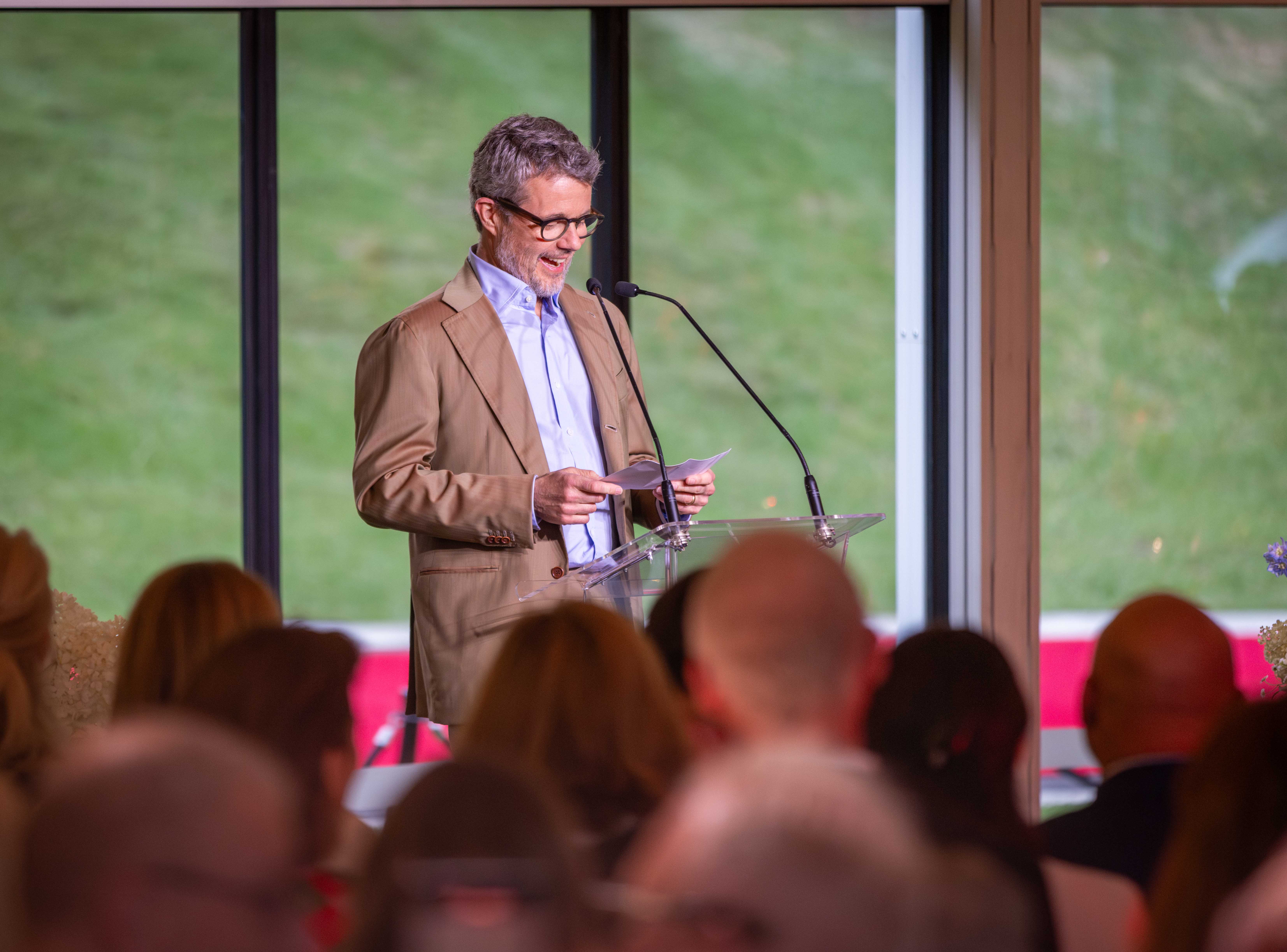 His Majesty The King of Denmark stands at a clear podium, speaking into a microphone at the Royal Botanic Gardens. He's dressed in a tan suit and light blue shirt, holding papers. An audience sits in front of him, with a large window and green scenery in the background.