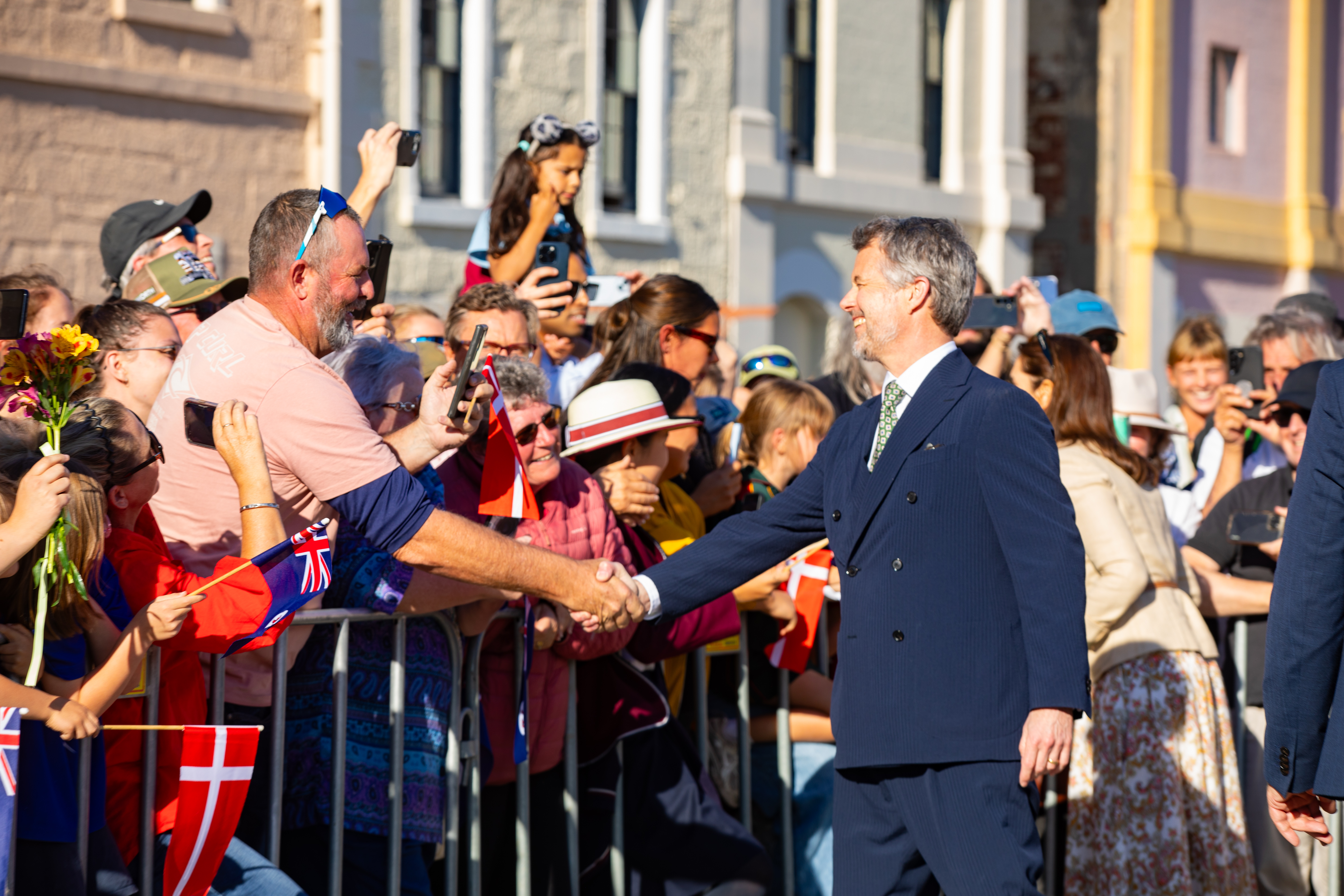 His Majesty The King of Denmark smiles, shaking hands with an onlooker over a metal barrier, while many people behind the barrier take photos with their phones or wave flags.