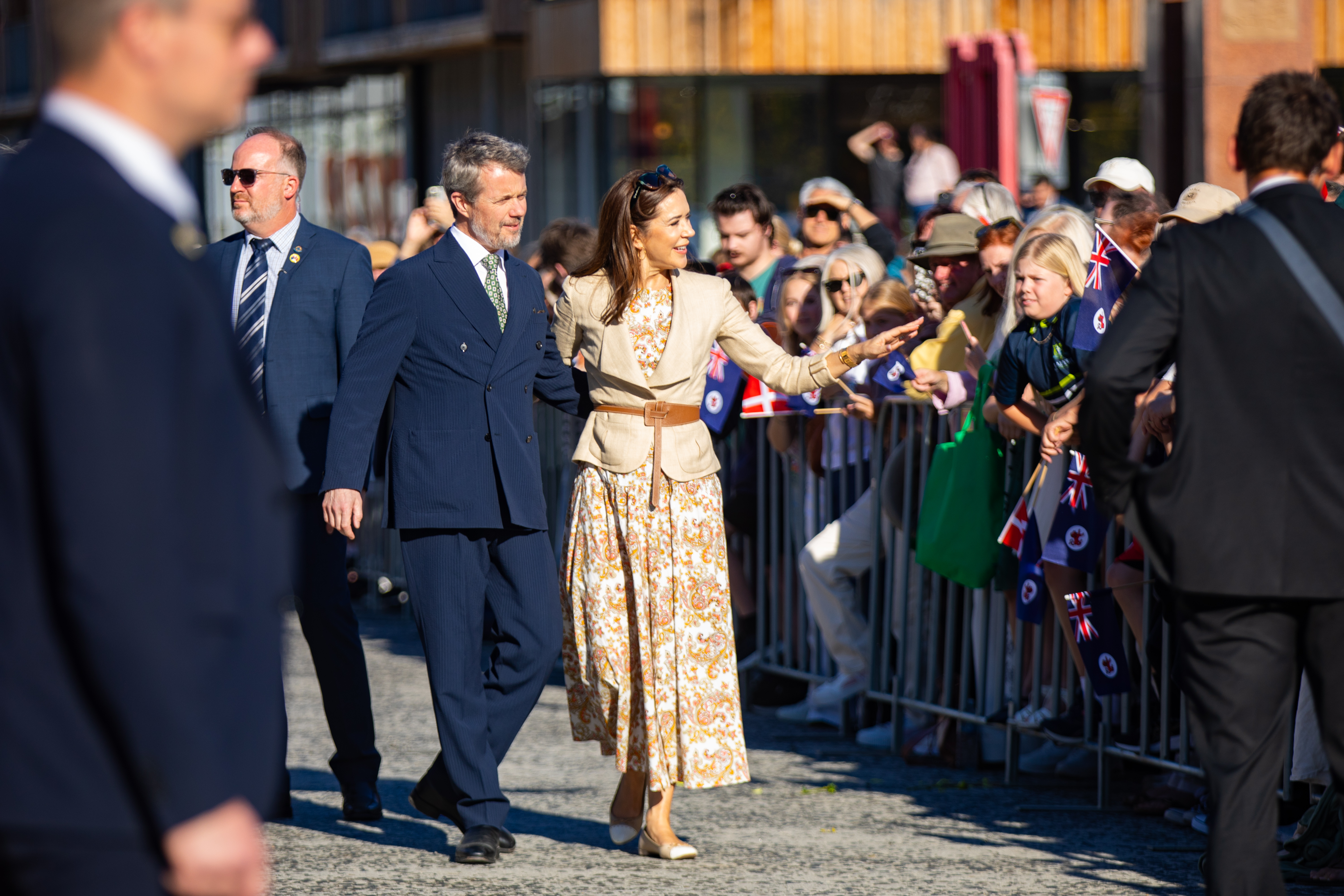 Her Majesty The Queen of Denmark in a tan blazer and floral skirt walks with His Majesty The King of Denmark, with a security detail and crowd nearby. Her Majesty points with her right hand as the crowd waves small flags.
