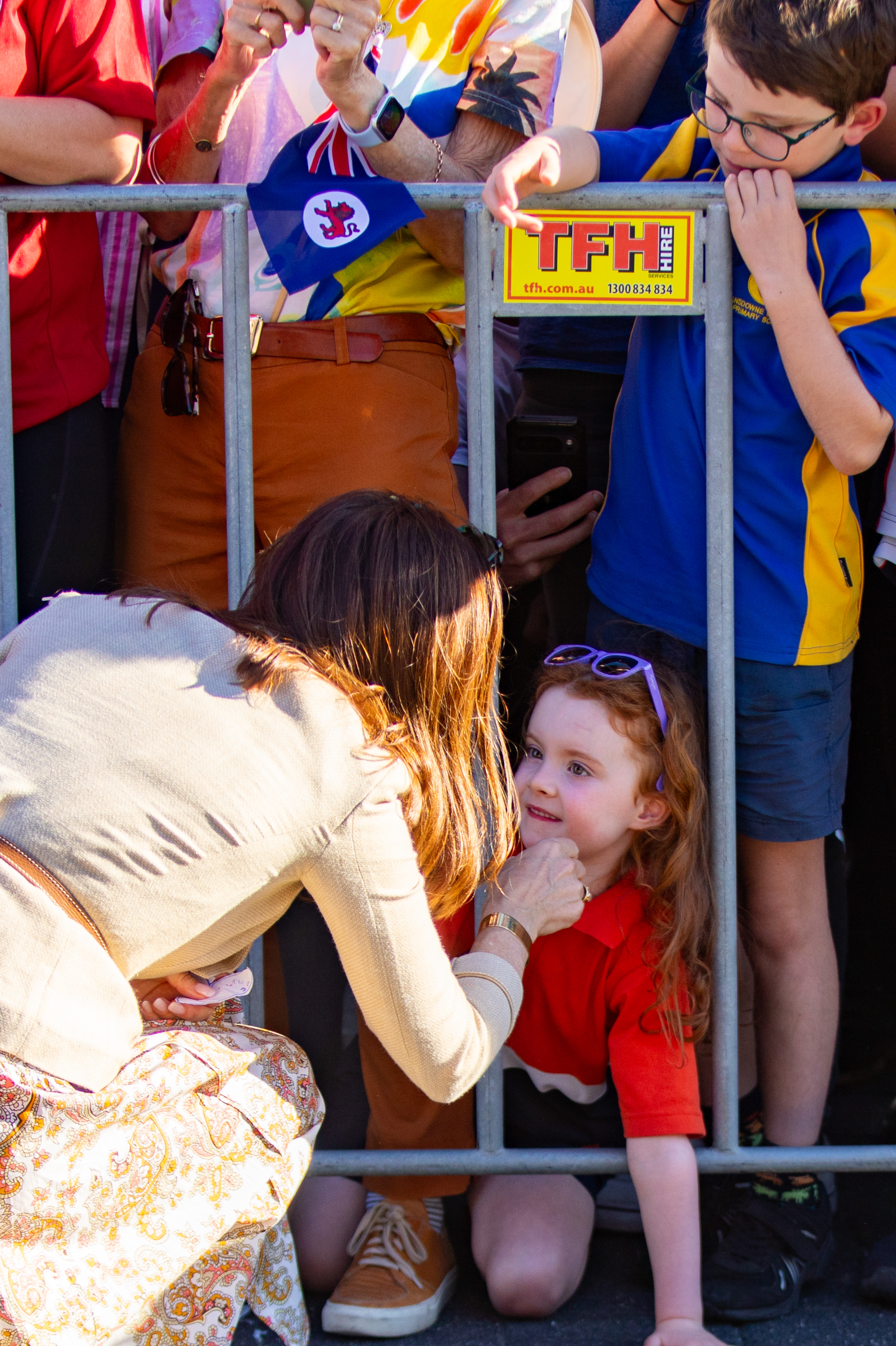 Her Majesty The Queen of Denmark in a floral skirt kneels by a metal fence, touching the face of a red-shirted girl with auburn hair. Behind the fence, a boy in blue and yellow school uniform peers over the top.