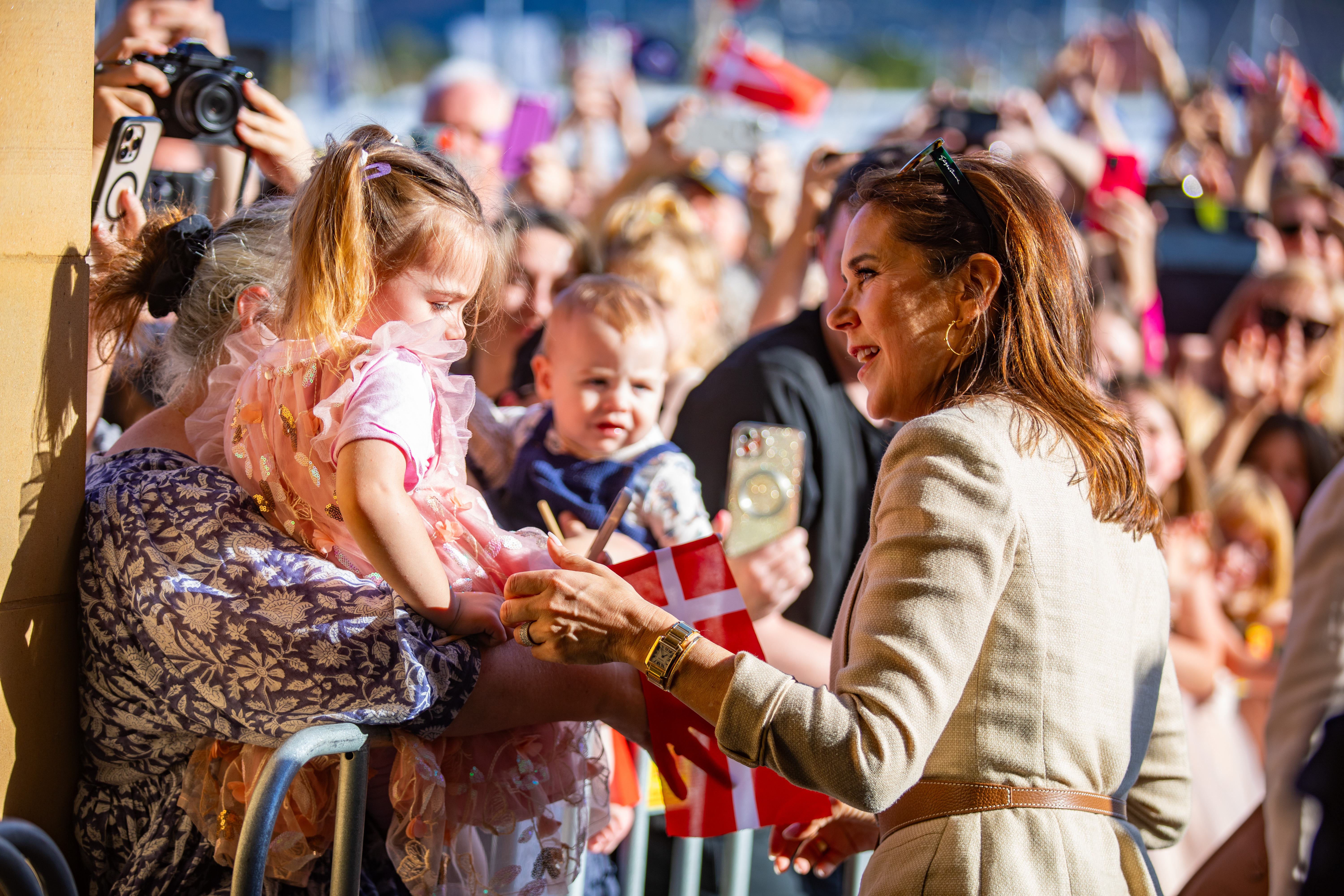 Amidst a crowd, Her Majesty The Queen of Denmark gestures to a child held by an adult. People behind them wave flags and hold up phones and cameras, capturing the moment in bright sunlight.