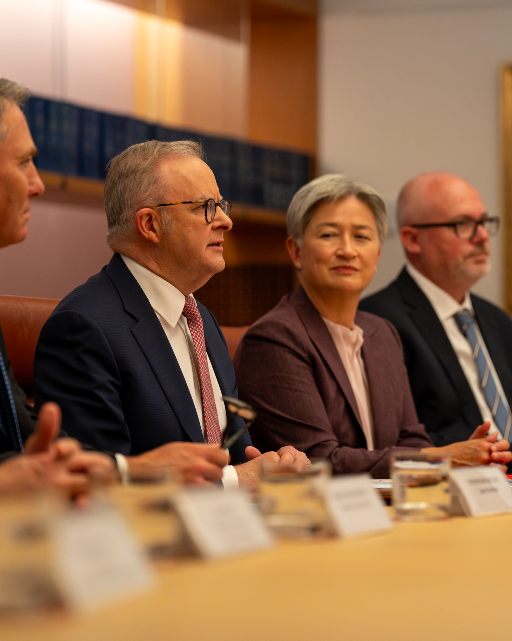 A group of officials sit at a long table in a meeting room, listening as Prime Minister Albanese speaks.