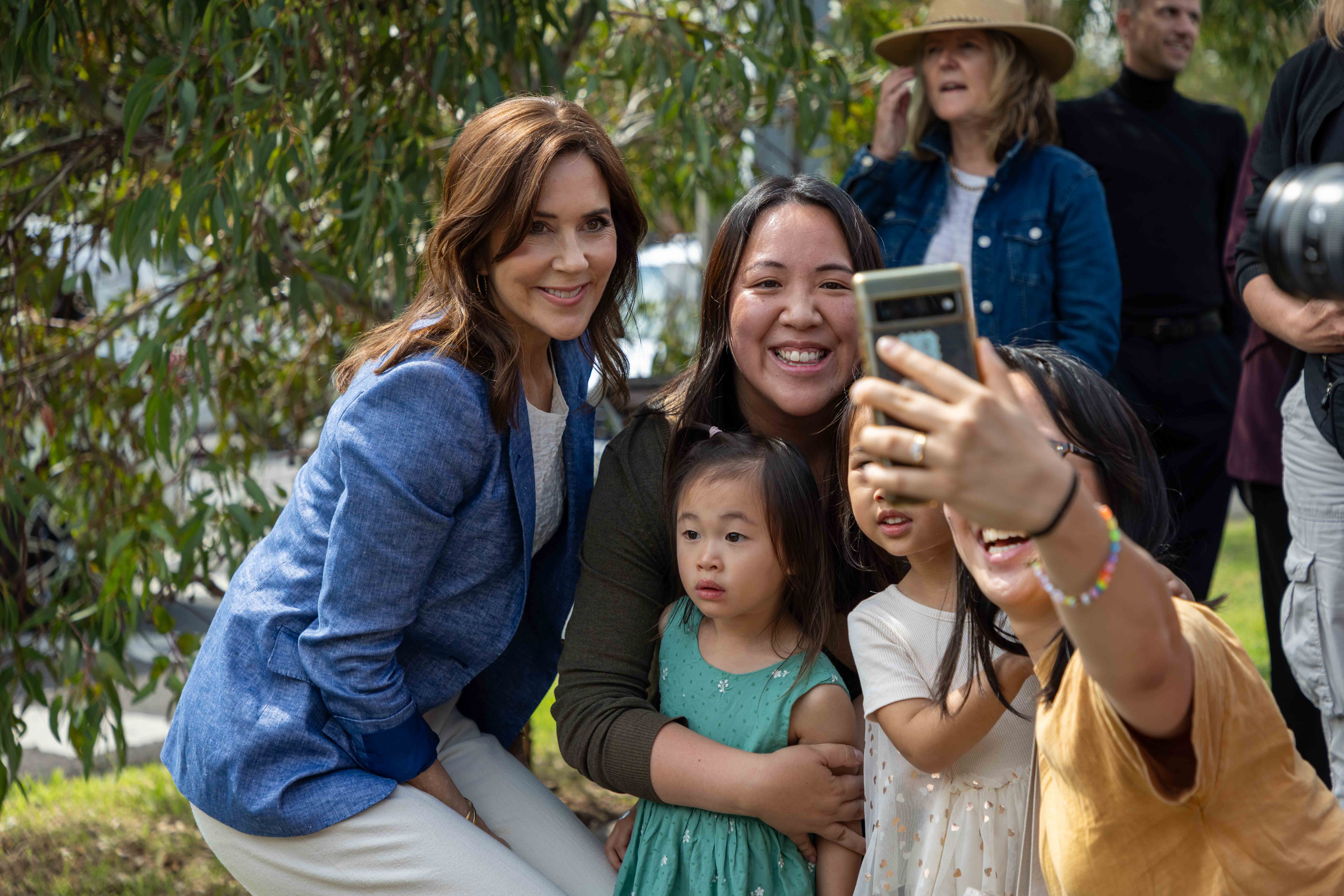 Her Majesty The Queen of Denmark poses in a selfie with a woman holding two little girls. An older girl in a yellow shirt holds the phone for the picture. In the background are several blurred adults.