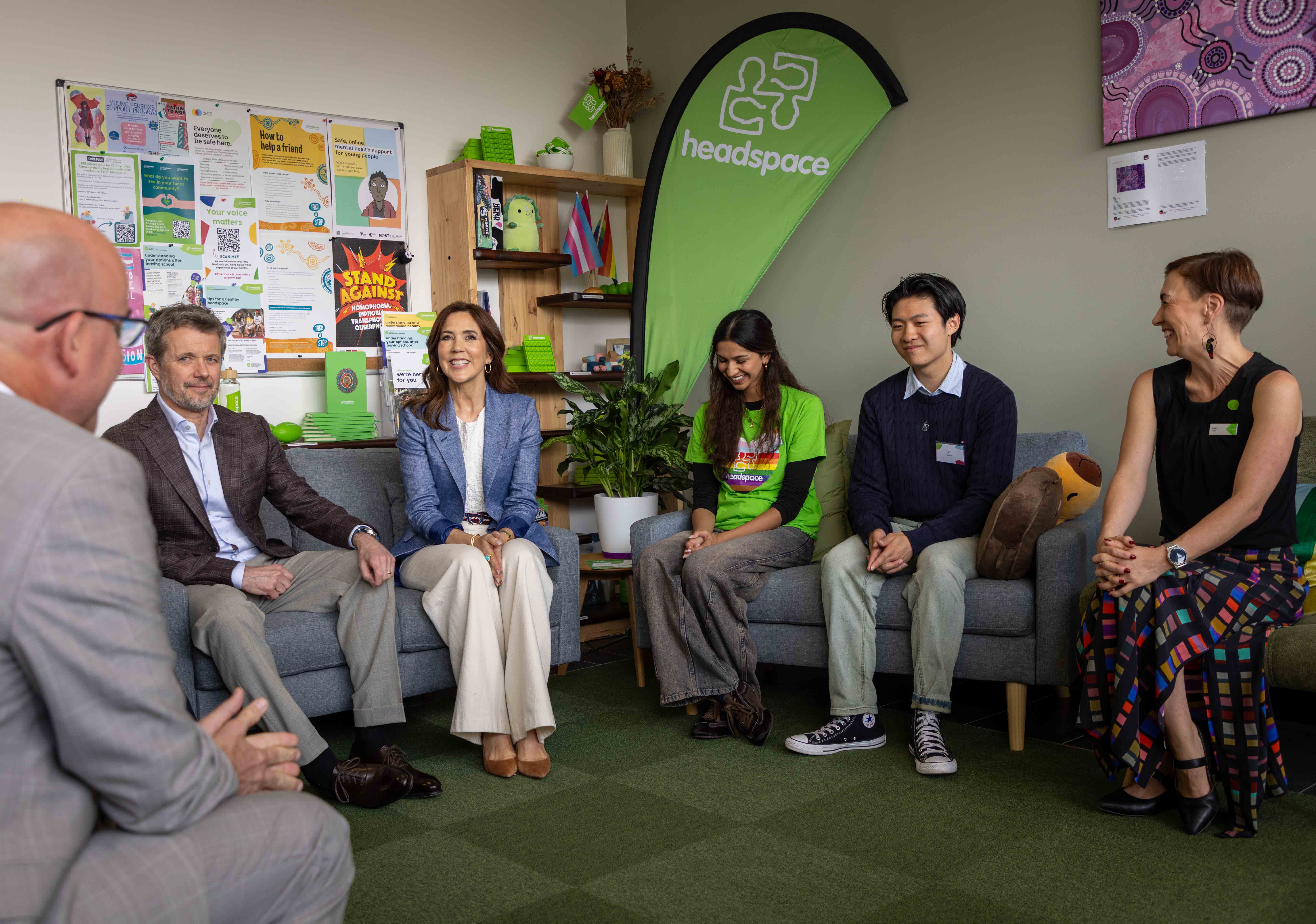 A group sits in a circle in a casual room. "headspace" is seen on a banner. Her Majesty The Queen of Denmark sits on a grey couch with two young adults. On a nearby couch, a person in grey suit sits opposite them. In background is a wall of posters.