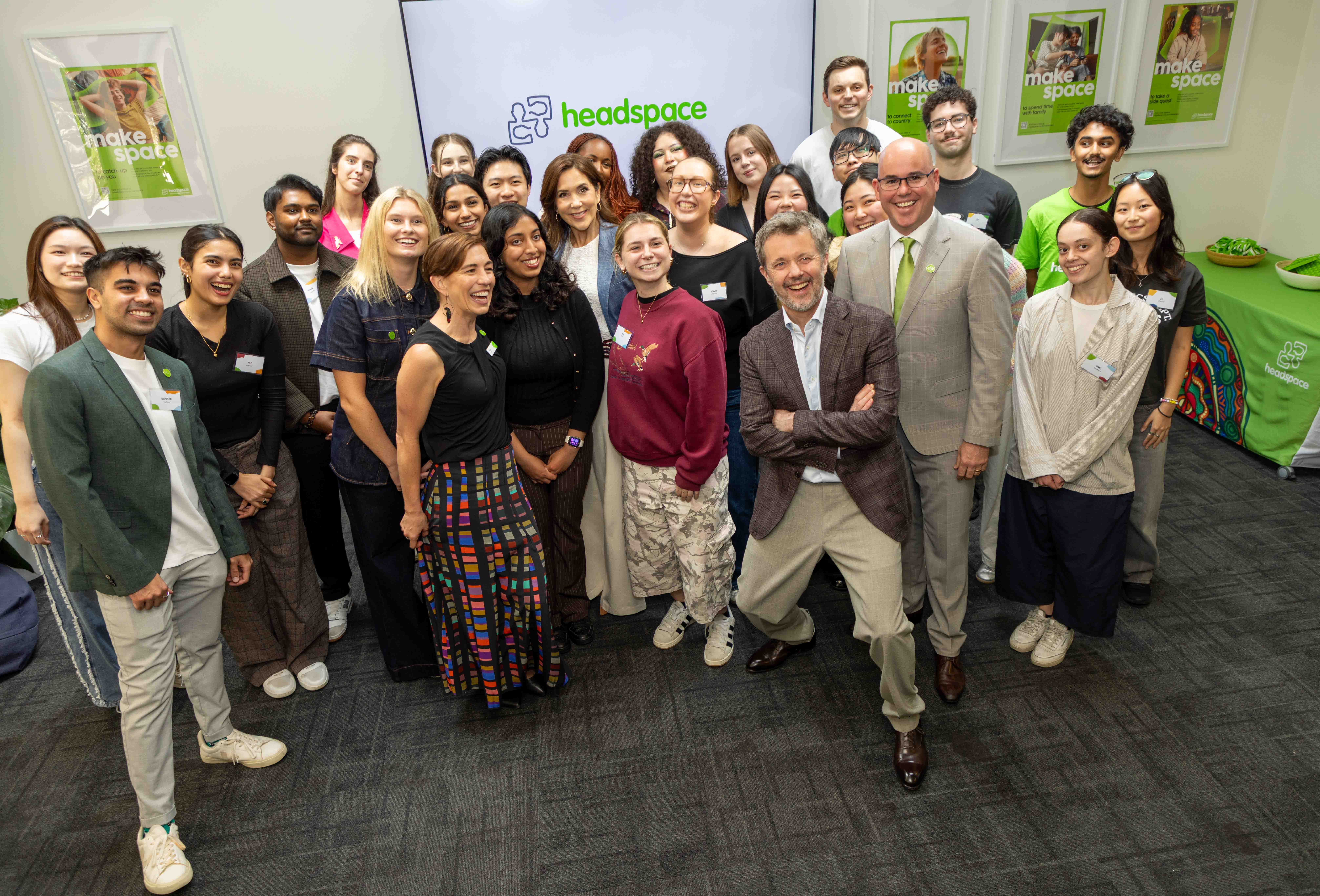 A group of about 30 people, diverse in race and gender, smile at the camera with His Majesty The King of Denmark in front of a screen with the text "headspace." Some hold name tags. Posters read "make space." 
