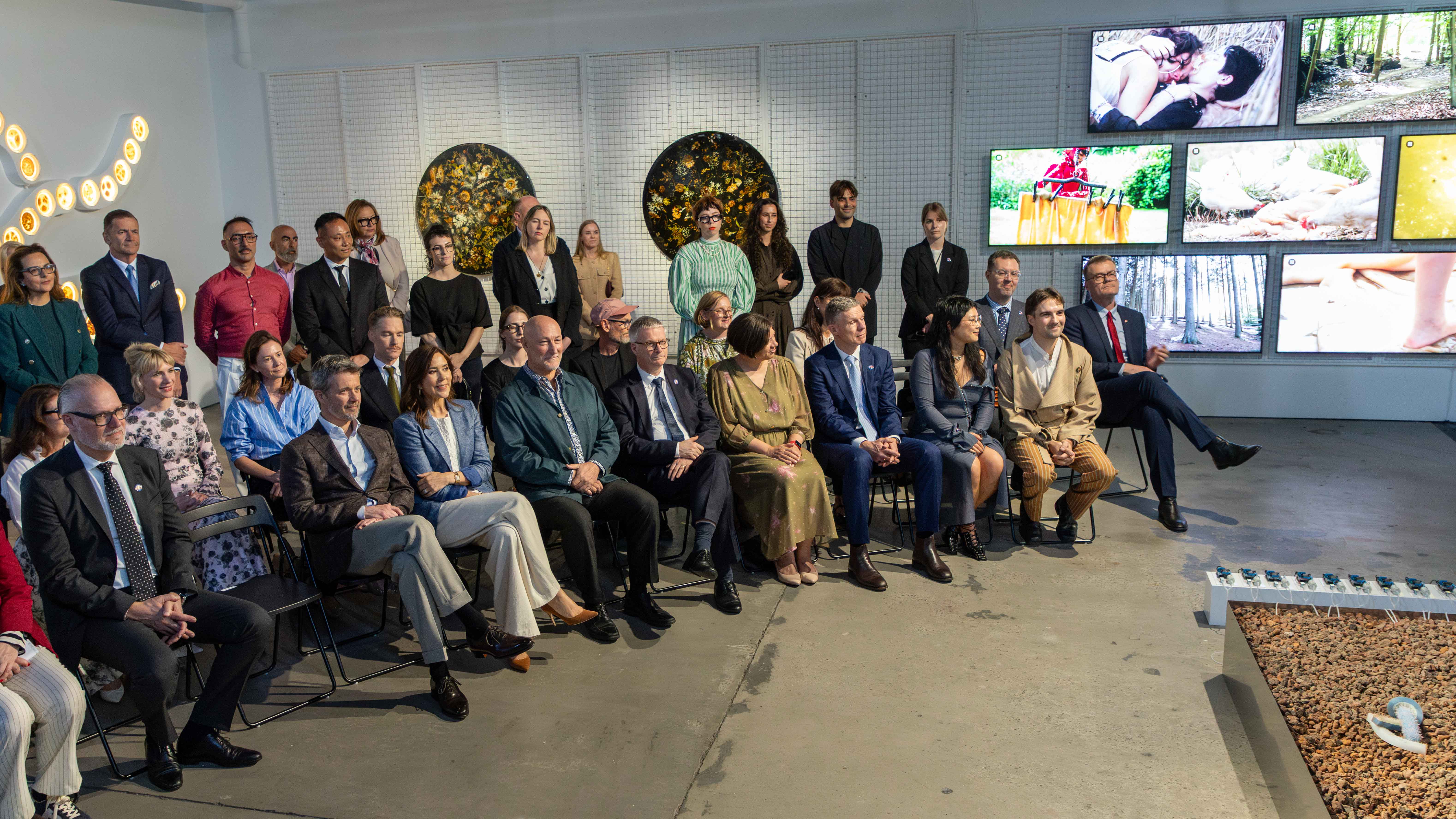 Group of people including Their Majesties The King and Queen of Denmark seated in rows, facing forward, at Art Bank, Melbourne, with artworks on the wall behind them. Includes lit wall art and video screens displaying scenes.