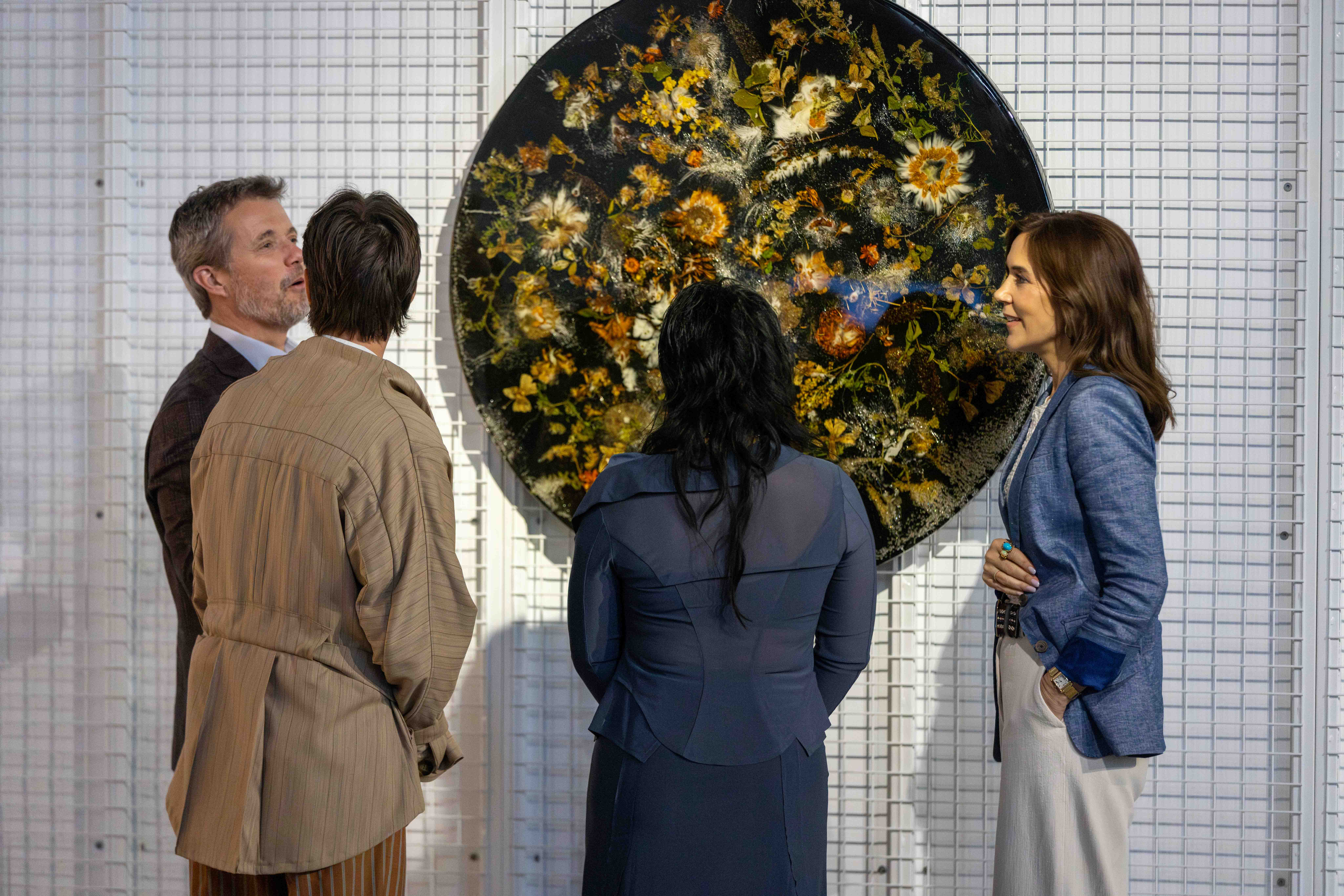Their Majesties The King and Queen of Denmark stand with two people in front of round floral artwork on a white grid wall. The floral art has a dark background with scattered yellow and white flowers.