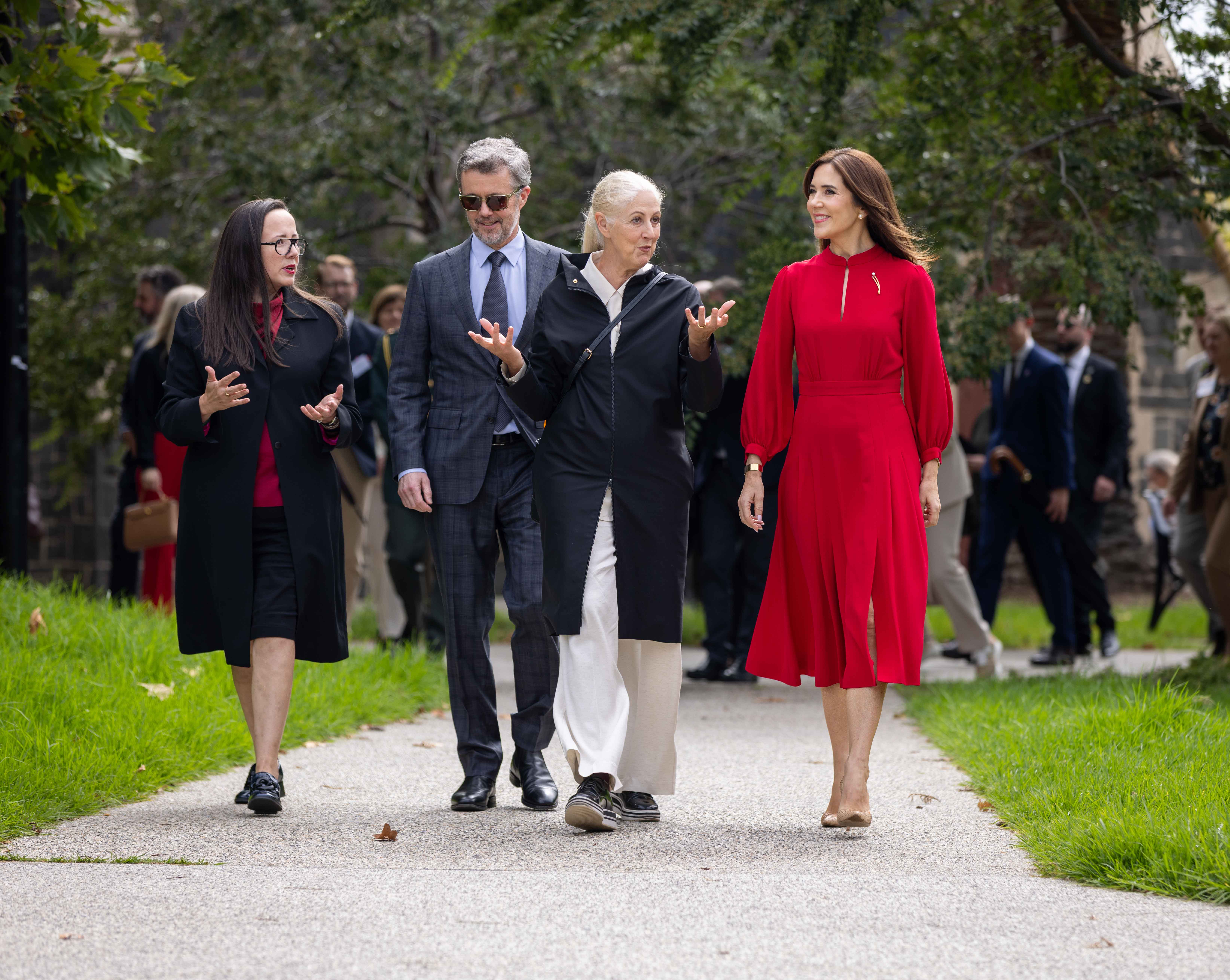 Four people walk on a path; Her Majesty The Queen of Denmark is in a red dress is on the right. The second person to the left is His Majesty The King of Denmark in a suit and sunglasses; a woman in a black coat is on the left of His Majesty and another woman in a black coat with white pants is next to Her Majesty.