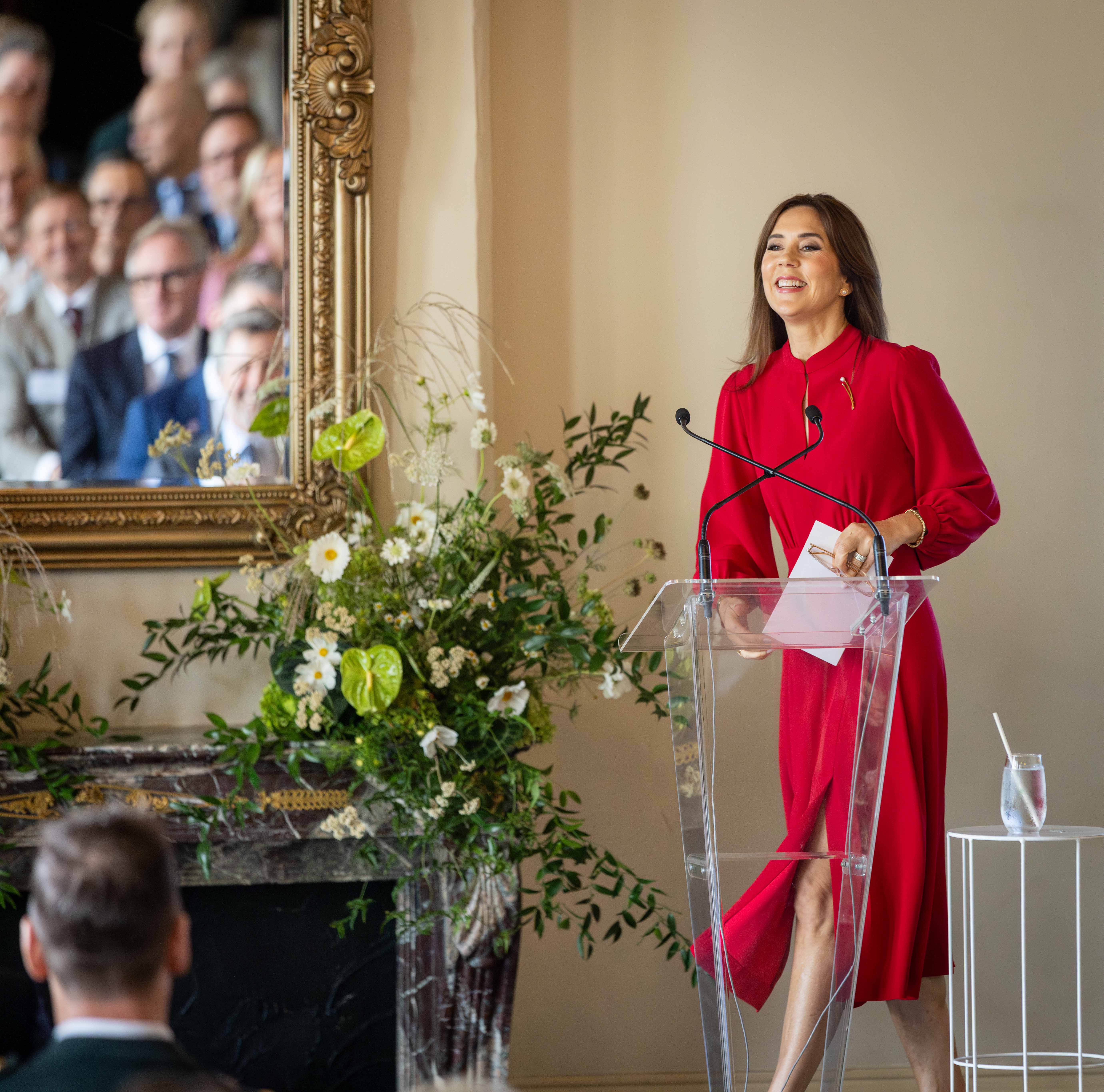 Her Majesty The Queen of Denmark in a red dress speaks at a clear podium. Behind her is an ornate mirror reflecting an audience. A glass of water with a straw sits on a small white table to her right. Flowers adorn the space to the left of the podium.