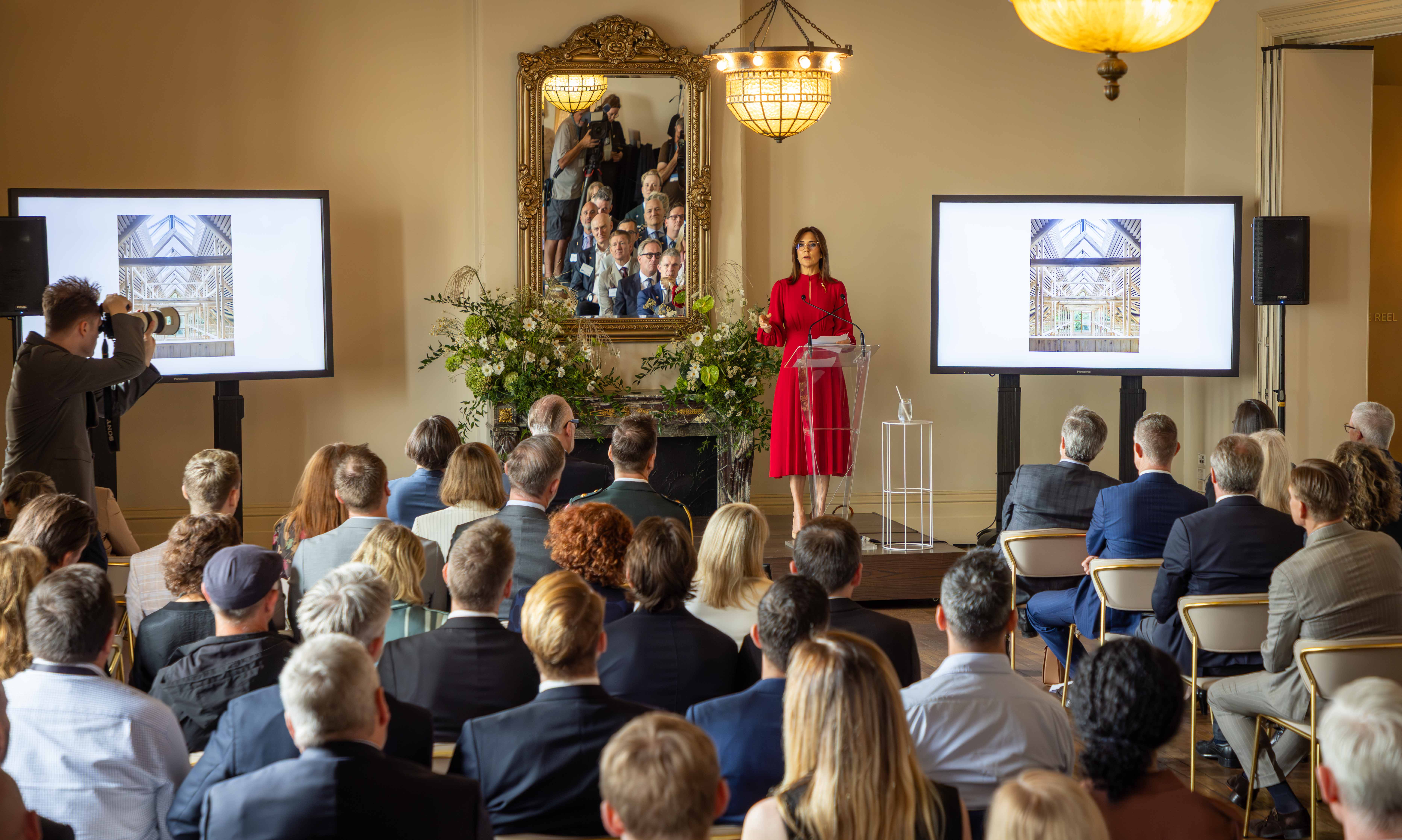 Her Majesty The Queen of Denmark stands at a podium giving a presentation to an audience. Two screens flank her with an image of a building under construction. A photographer takes photos to the left of the frame.