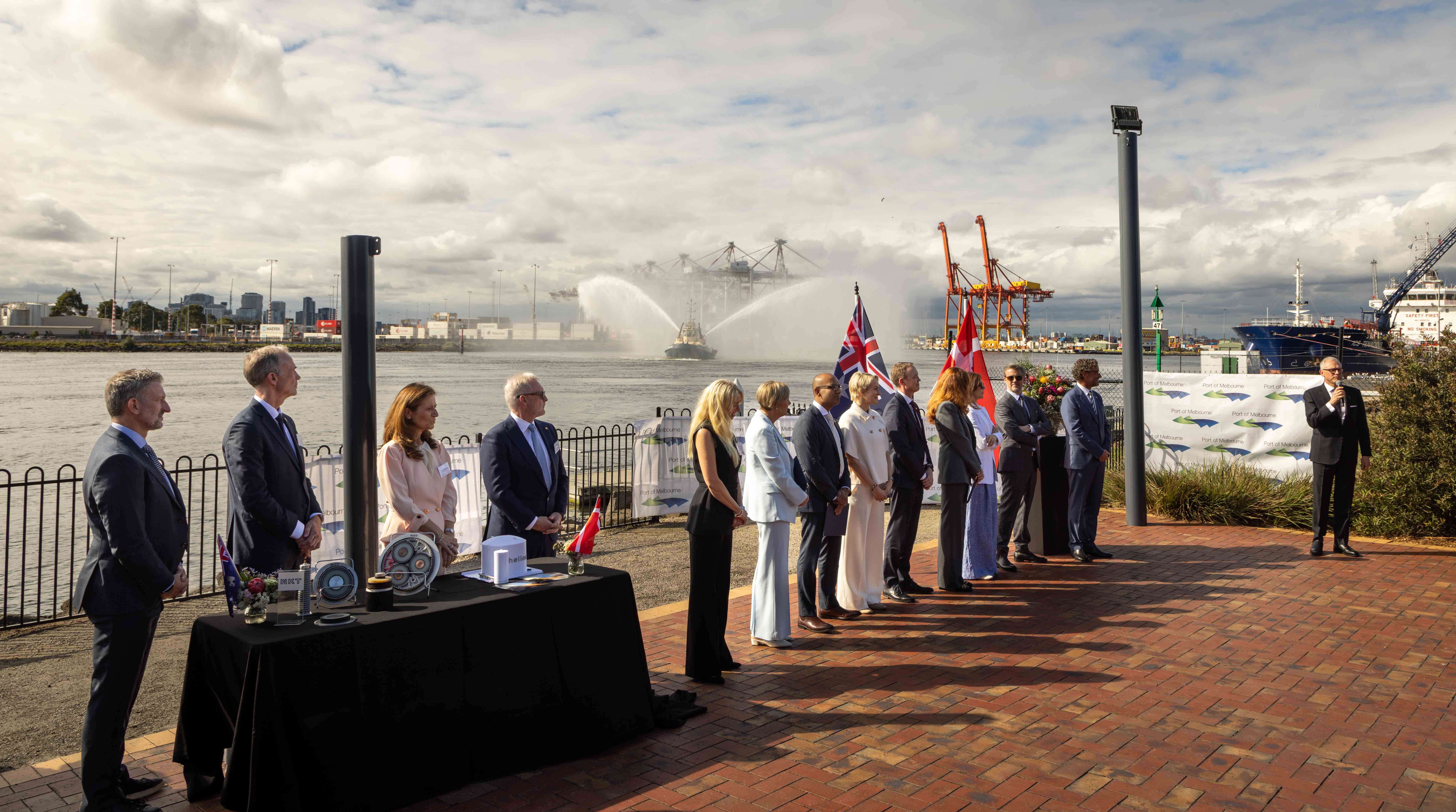 Their Majesties The King and Queen of Denmark attend an engagement at Port of Melbourne. A waterside ceremony shows a line of dignitaries, including a speaker at right, with the text "Port of Melbourne" on a banner behind them. Cranes and a ship spraying water feature in the background.