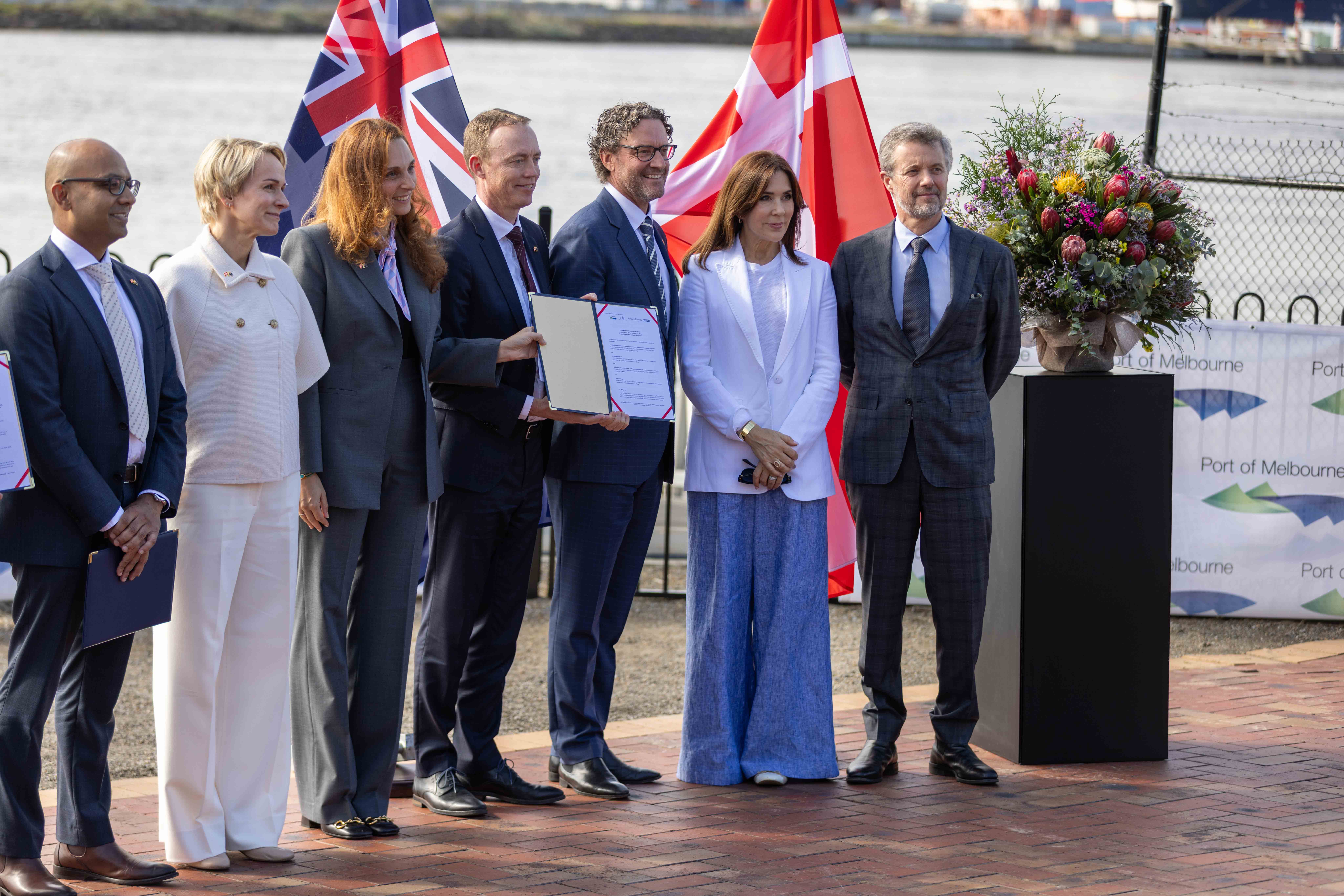 A group of eight people including Their Majesties The King and Queen of Denmark stand outdoors in formal attire, holding documents against a backdrop of UK and Danish flags. Text visible: "Port of Melbourne".
