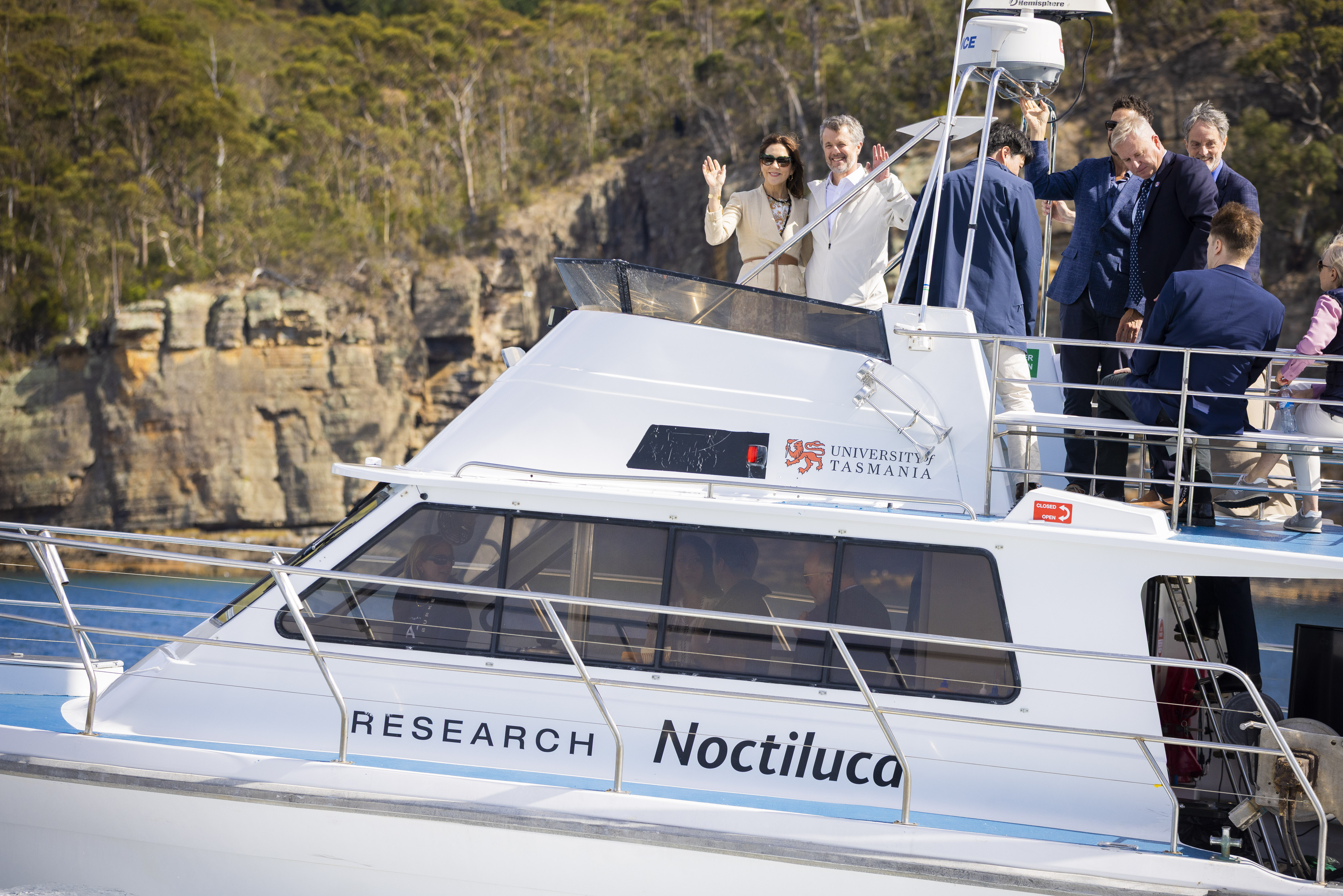 People on a 'RESEARCH Noctiluca' boat, with 'UNIVERSITY of TASMANIA' logo. Their Majesties The King and Queen of Denmark wave from the boat's front. Other people are standing on the boat.