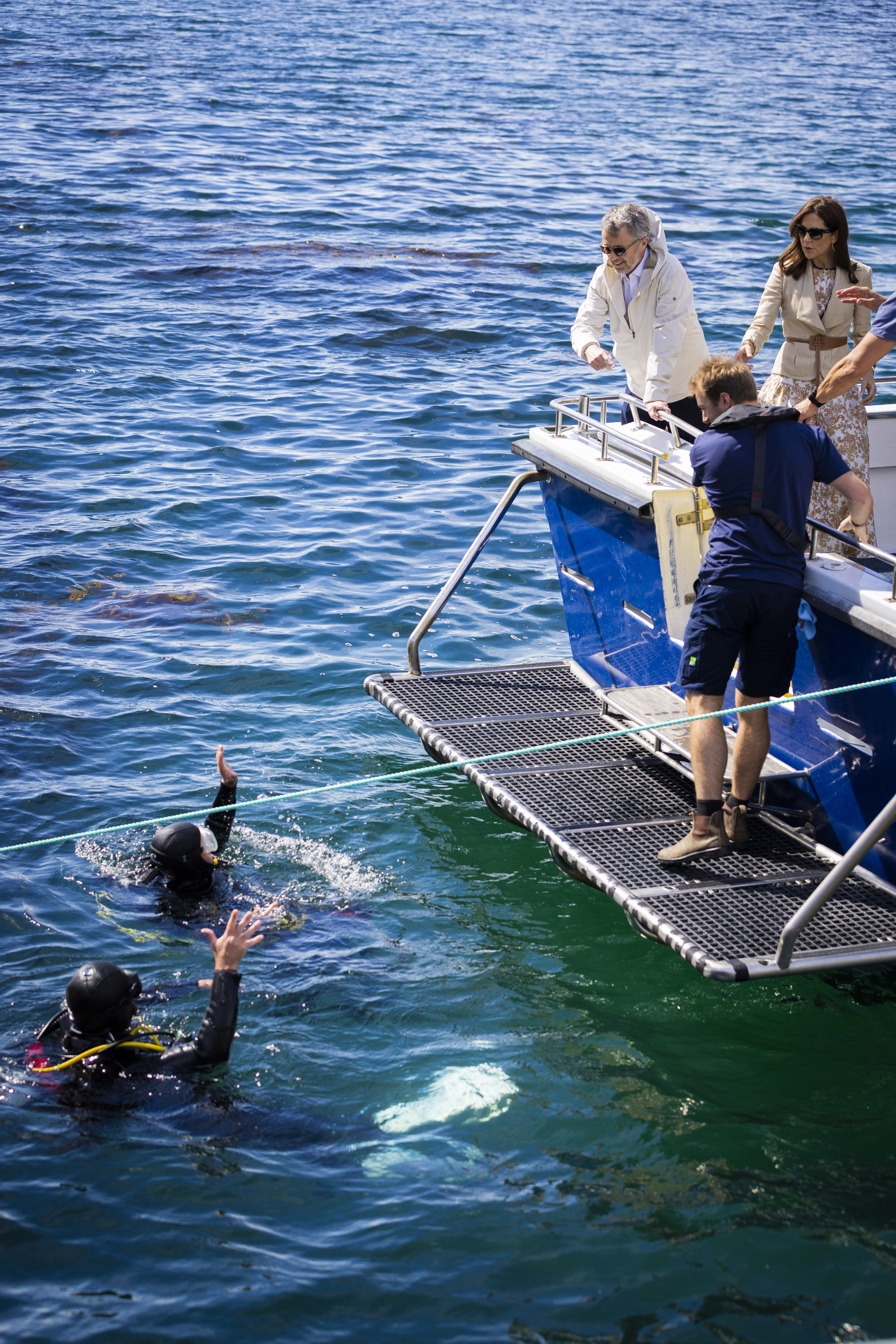 Ocean scene: divers with raised hands in blue water by a boat. On the boat's platform, Their Majesties The King and Queen of Denmark observing from the deck above. The boat is blue and white with a metal platform.