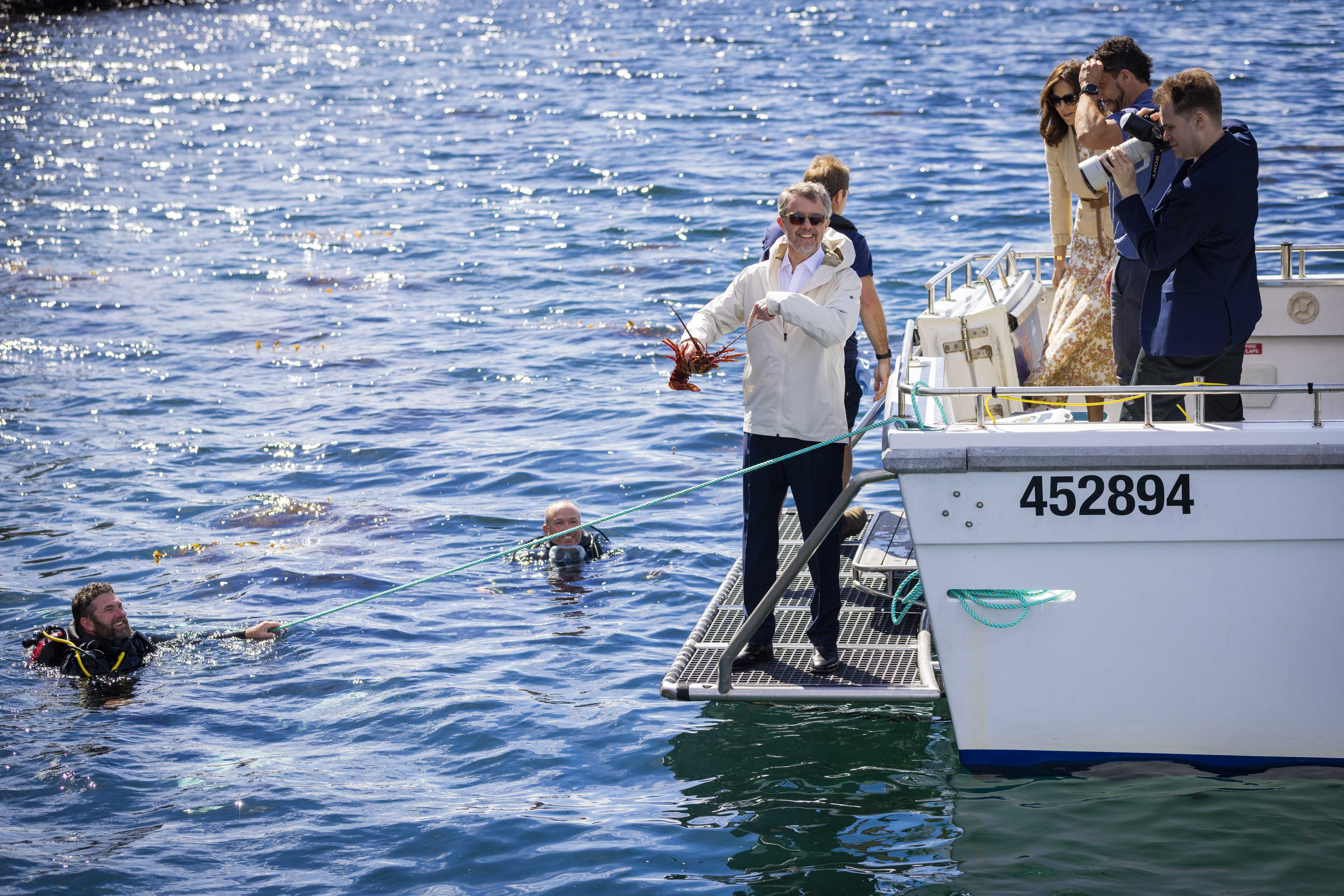 On a sunny day, His Majesty The King of Denmark on a boat ladder holds a lobster as two scuba divers emerge from the water. The boat displays the number 452894. Several people stand on the deck, one taking pictures.