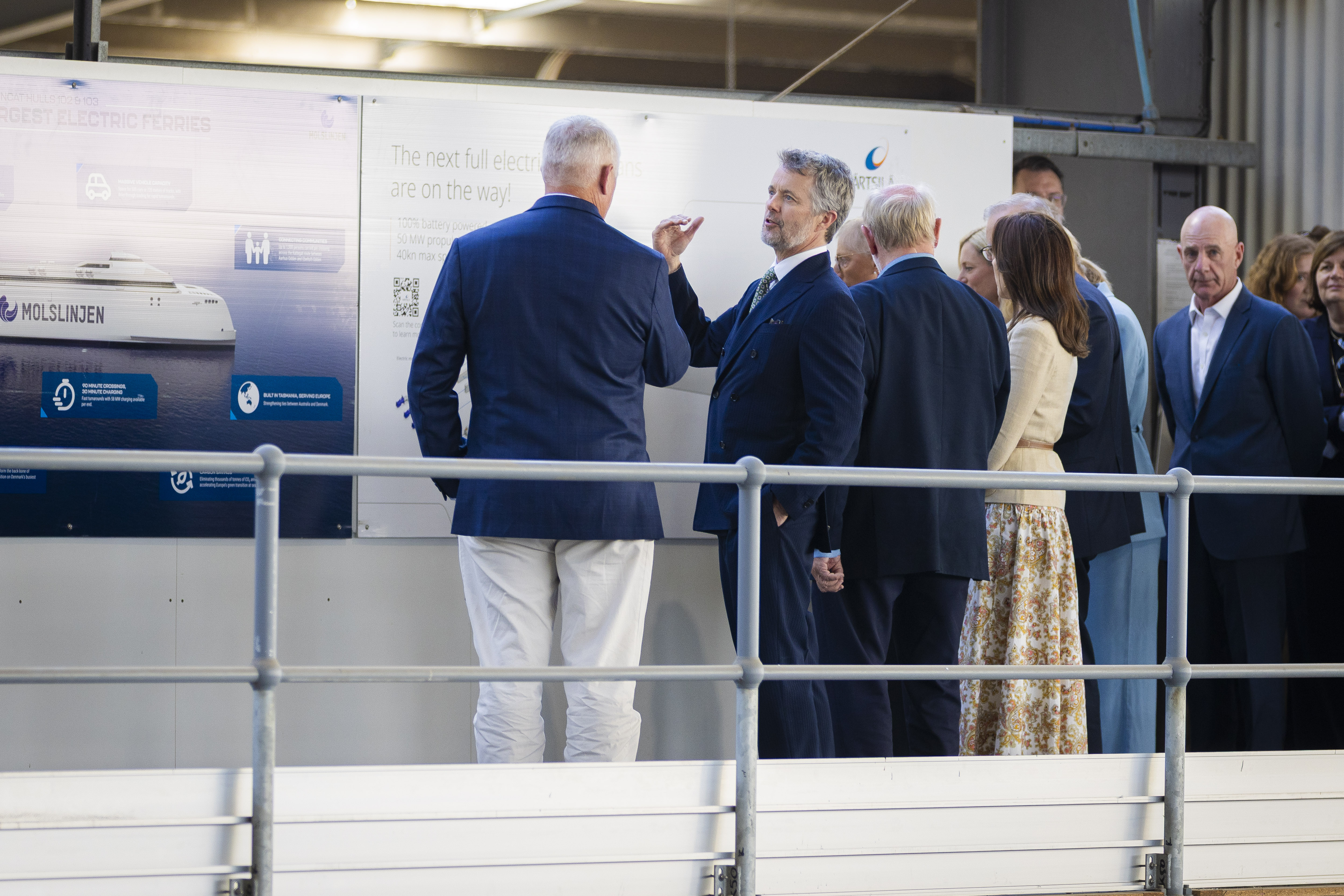 A group with His Majesty The King of Denmark stands by a poster that reads "The next full electric ferries are on the way! 100% battery powered, 50 MW propulsion, 40kn max speed". Text on another poster reads "LARGEST ELECTRIC FERRIES MOLSLINJEN". They are at INCAT Tasmania.