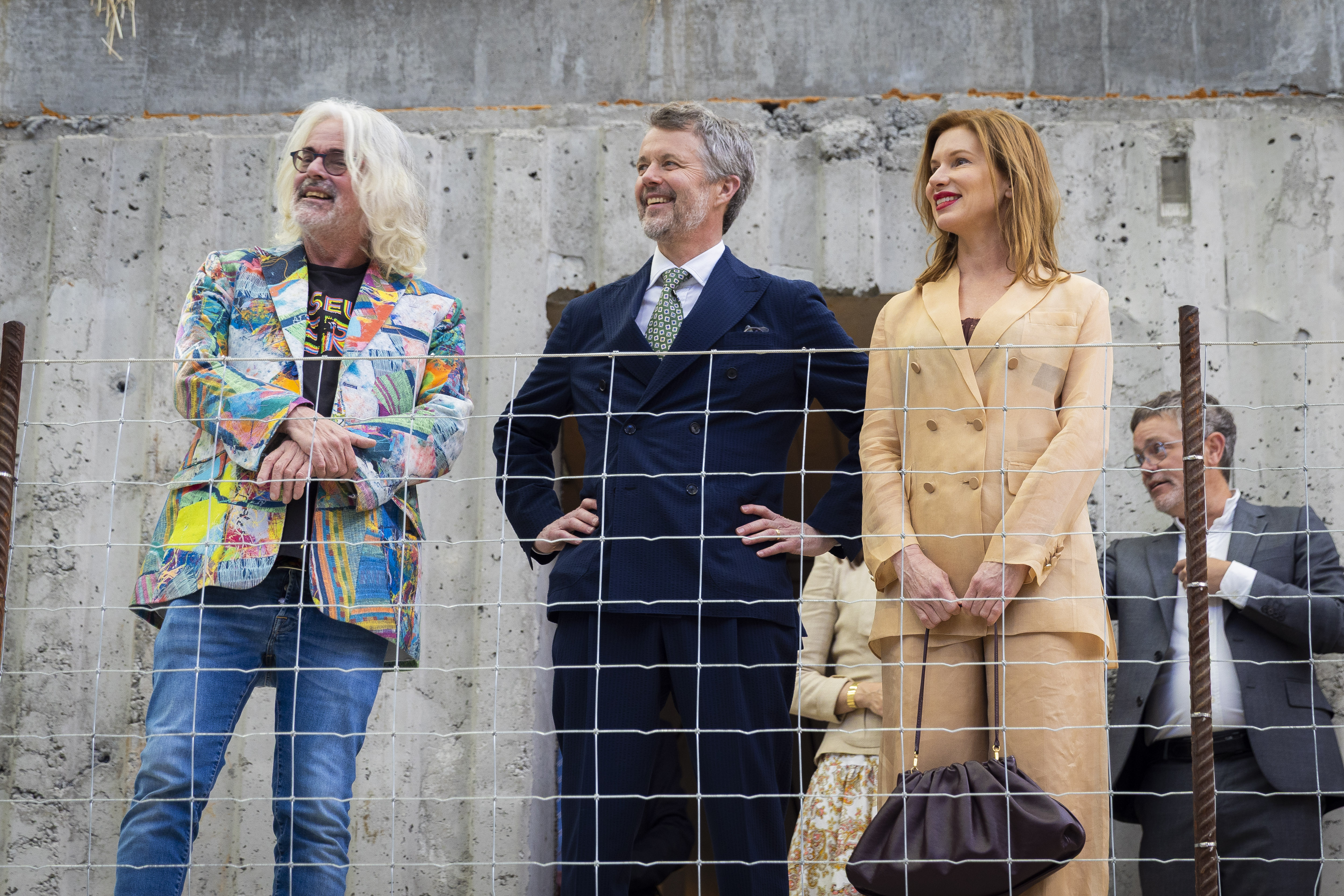 Four people including His Majesty The King of Denmark stand behind a wire fence with a gray concrete wall in the background at the Museum of Old and New Art. The first person wears a colorful blazer and jeans, His Majesty The King of Denmark wears a dark suit and green tie, and the third, a woman, wears a tan suit. The fourth person is in the background.