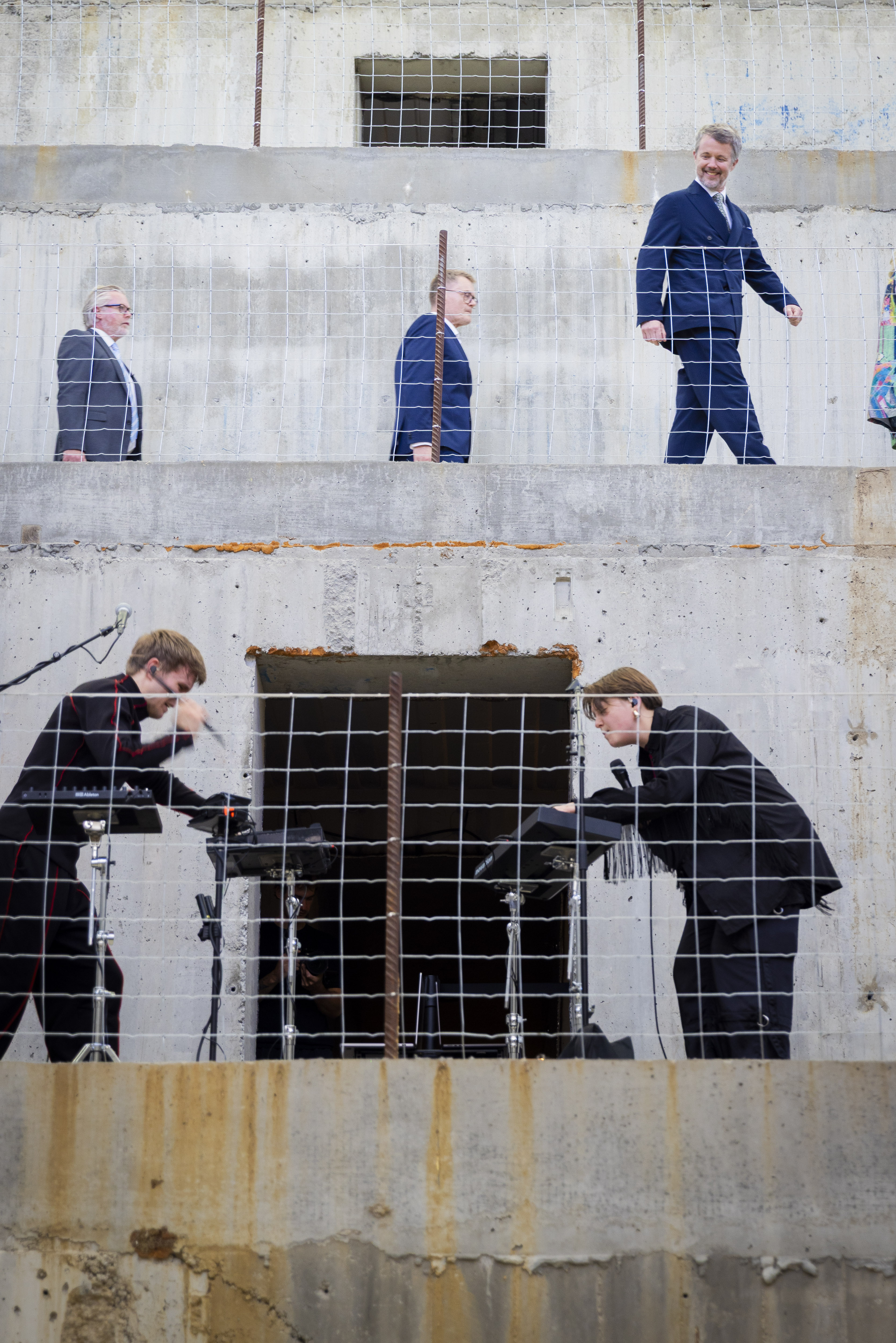 Eye-level shot of two people performing music by the band ROYA on electronic instruments behind a wire fence at the Museum of Old and New Art, with His Majesty The King of Denmark walking behind them, watching and smiling on higher ground against a gray concrete wall with square openings.