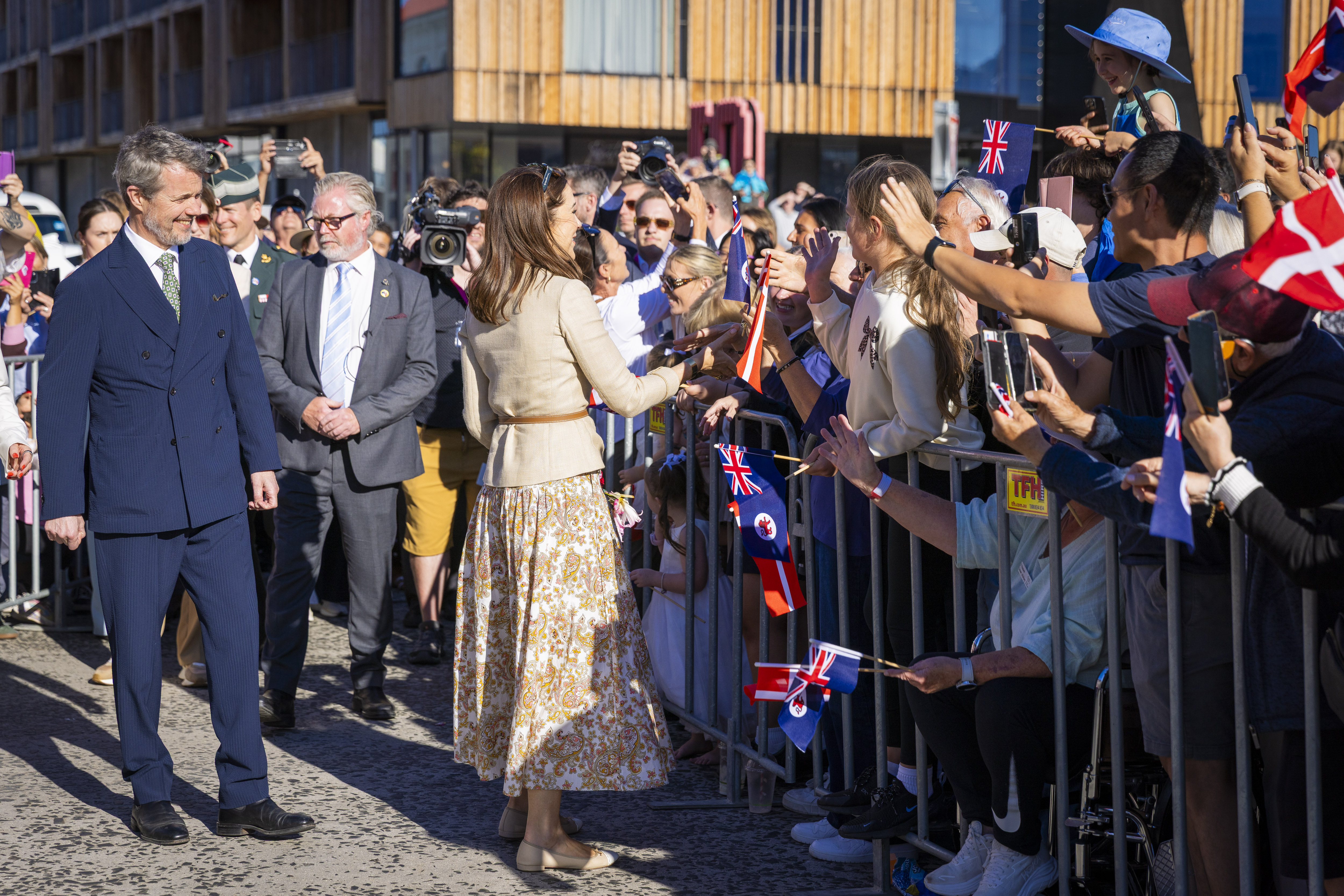 Their Majesties The King and Queen of Denmark greet onlookers behind a metal barricade. Many people hold up phones and flags while trying to get the couple's attention.
