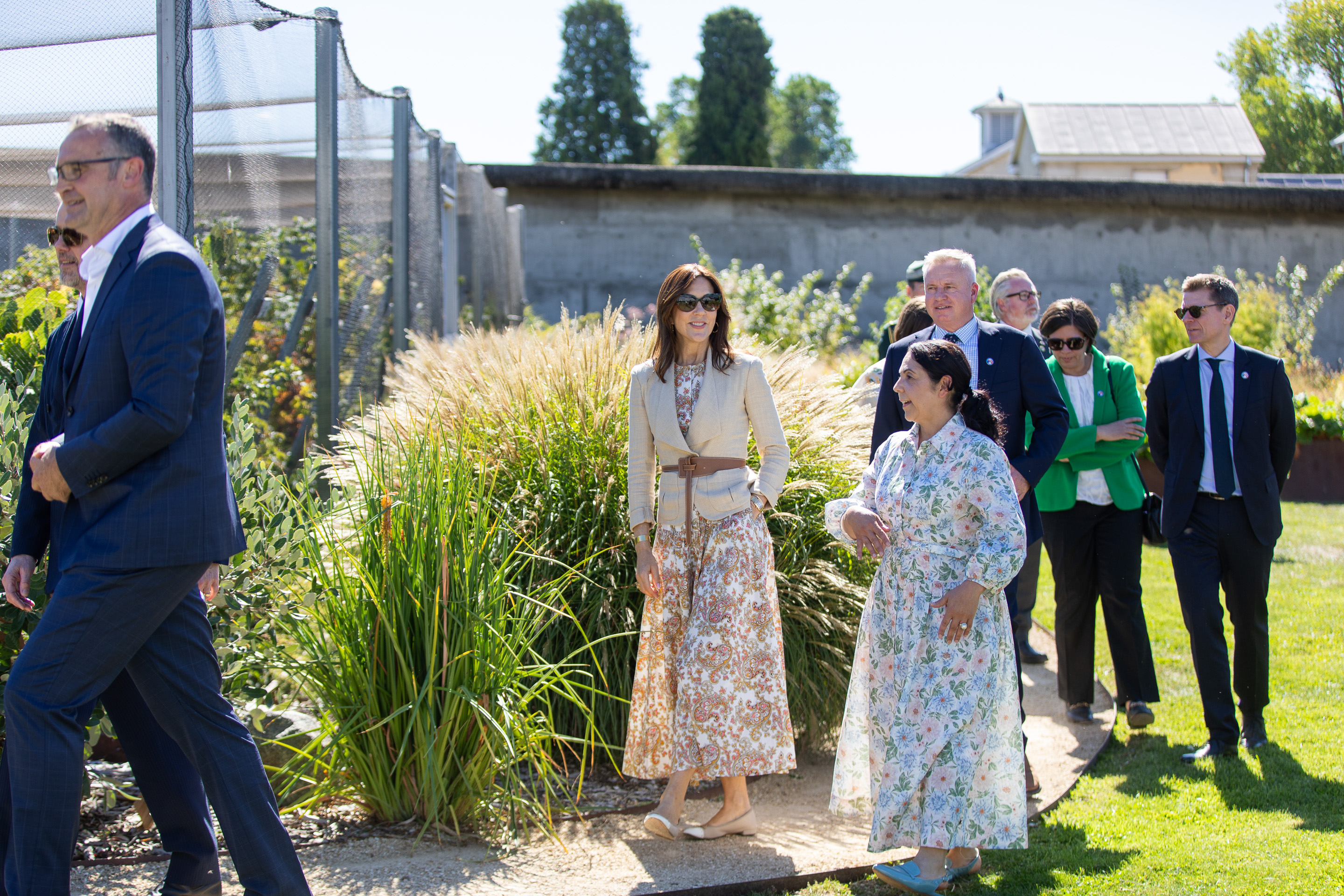 The image shows Her Majesty The Queen of Denmark accompanied by a group of people including men in suits and a woman in a green blazer, as they walk through a garden at the Agrarian Kitchen. Her Majesty is wearing a paisley dress and a tan jacket.