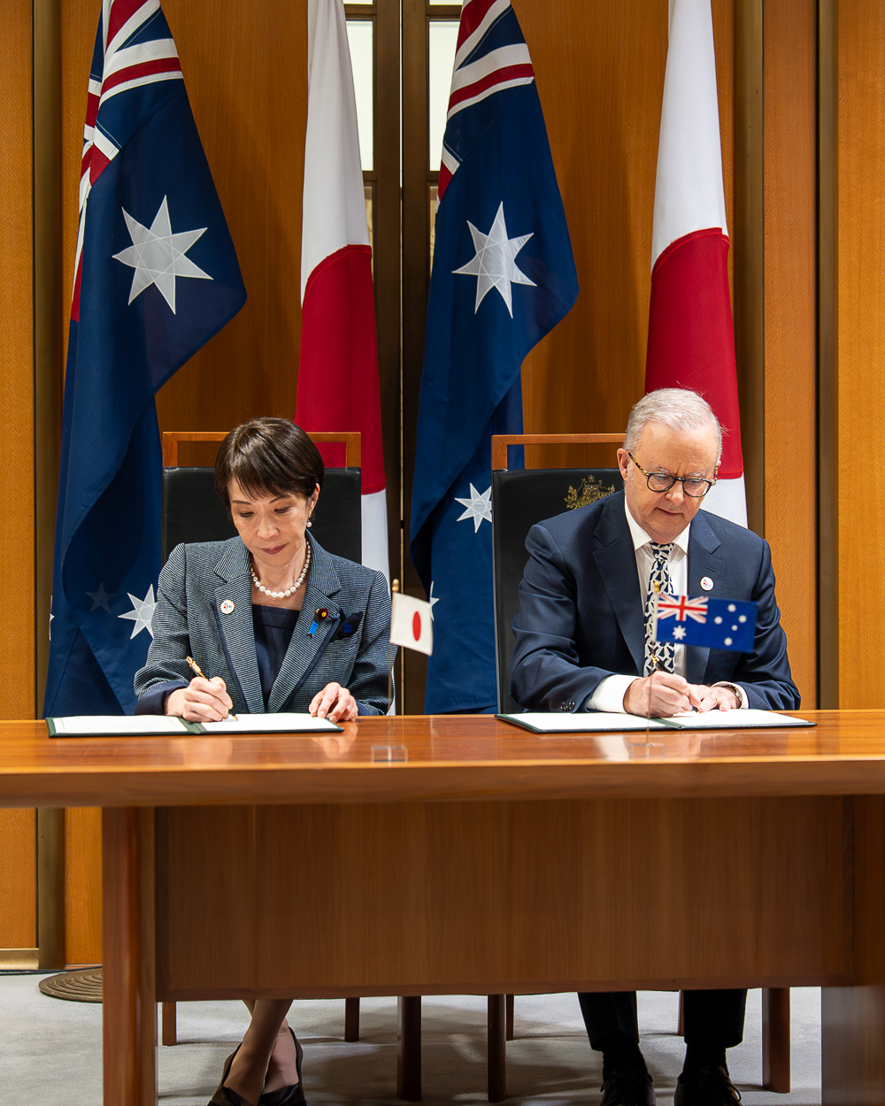Prime Minister Anthony Albanese and Prime Minister of Japan Sanae Takaichi sign documents at a formal bilateral meeting table with Australian and Japanese flags behind them.