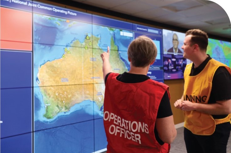 NEMA staff, dressed in operations officer vests, stand next to and point at a wall of screens. These screens display a map of Australia on the National Joint Common Operating Picture (NJCOP).