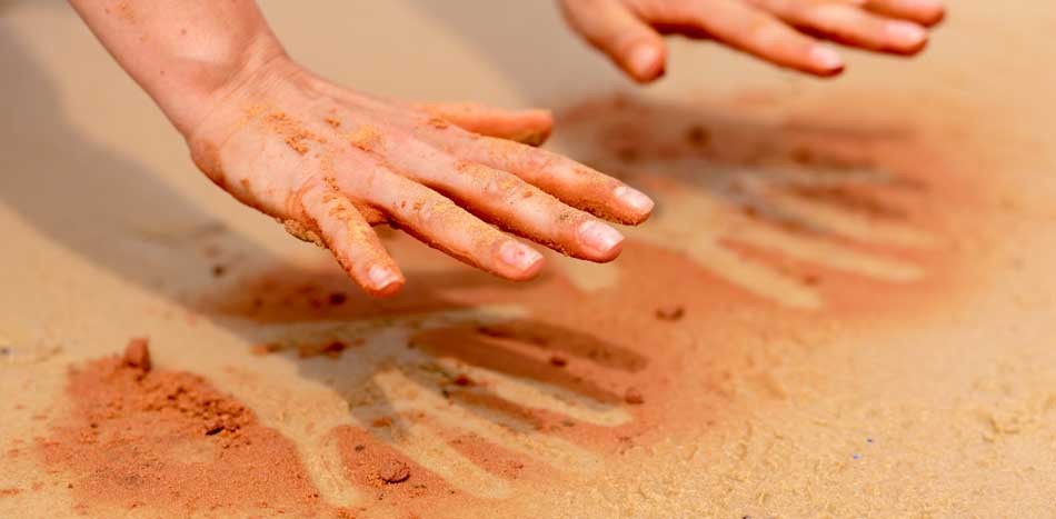 A pair of hands held above outlines of their own shape made from red sand