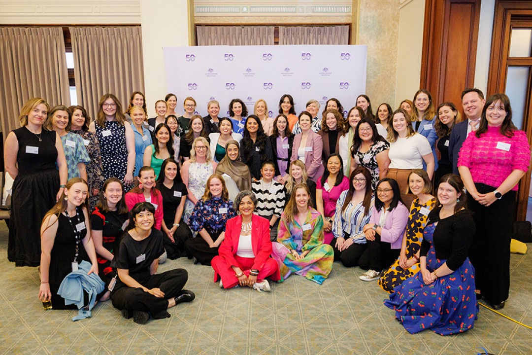 A group of 48 people pose for a group photo in front of a large purple banner printed with ‘Office for Women 50th Anniversary'.