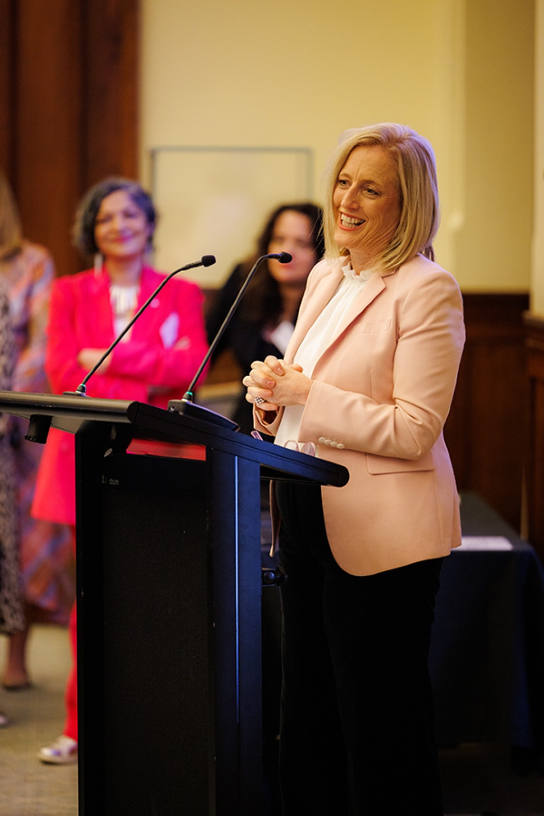 Senator the Hon Katy Gallagher, Minister for Women in a pale pink blazer stands behind a lectern, smiling while giving a speech.