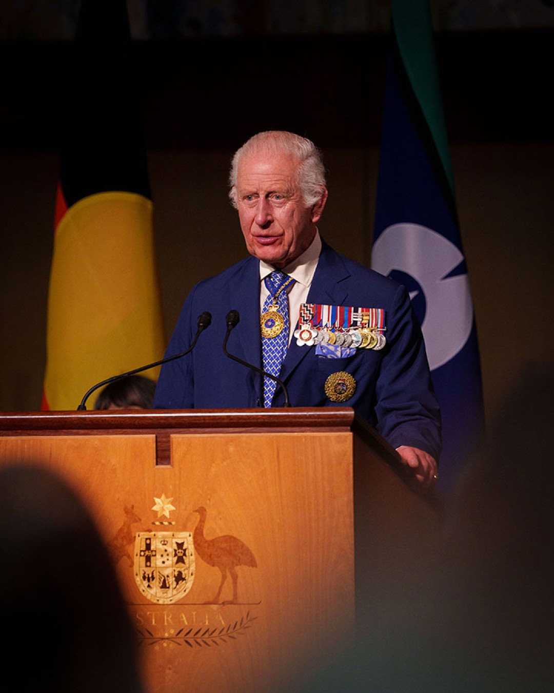 His Majesty stands at a lectern in front of the Australian Aboriginal Flag and the Torres Strait Islander Flag. The wooden lectern is inlayed with the Commonwealth Coat of Arms.