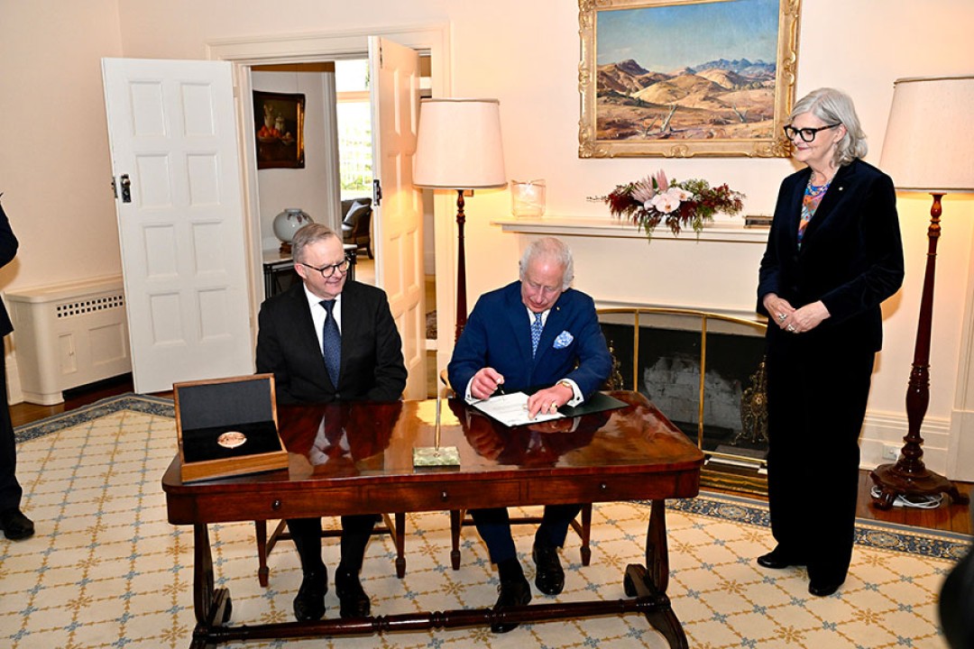 His Majesty signs a Royal Warrant. He is sitting at a desk next to Prime Minister Anthony Albanese while Her Excellency the Hon Sam Mostyn AC looks on. The new Great Seal of Australia sits in a case on the desk.